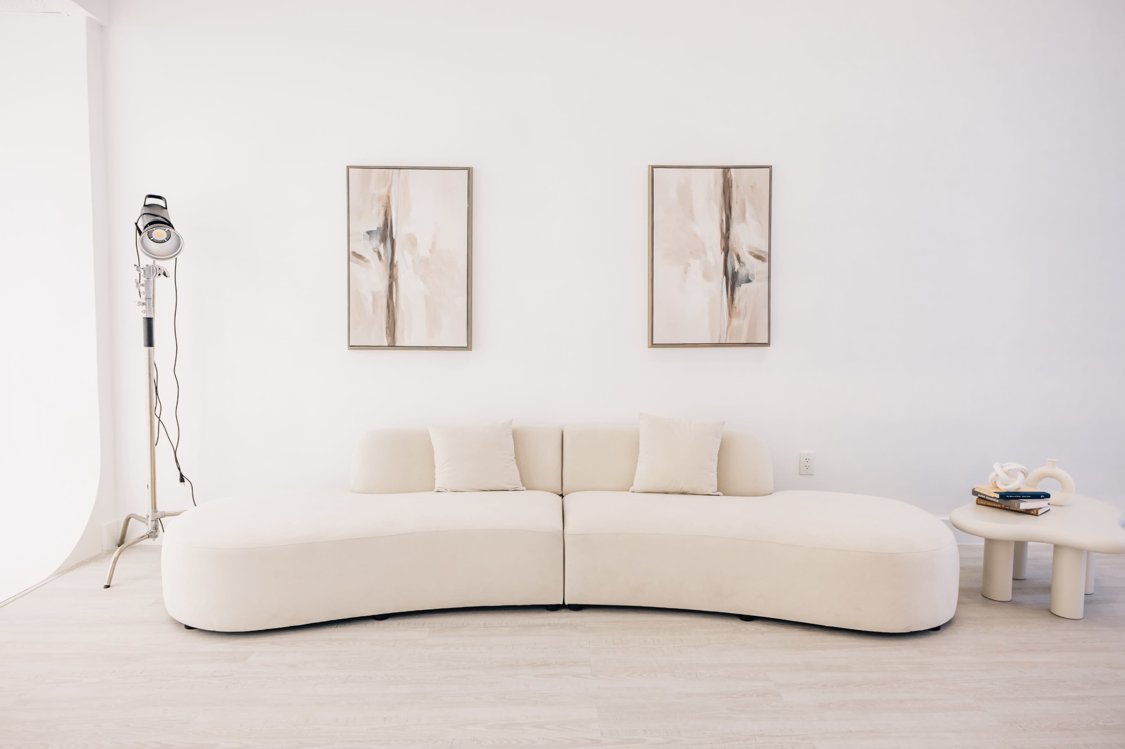 A curved beige sofa with two pillows sits against a white wall, flanked by two abstract paintings and a small side table with decorative items.