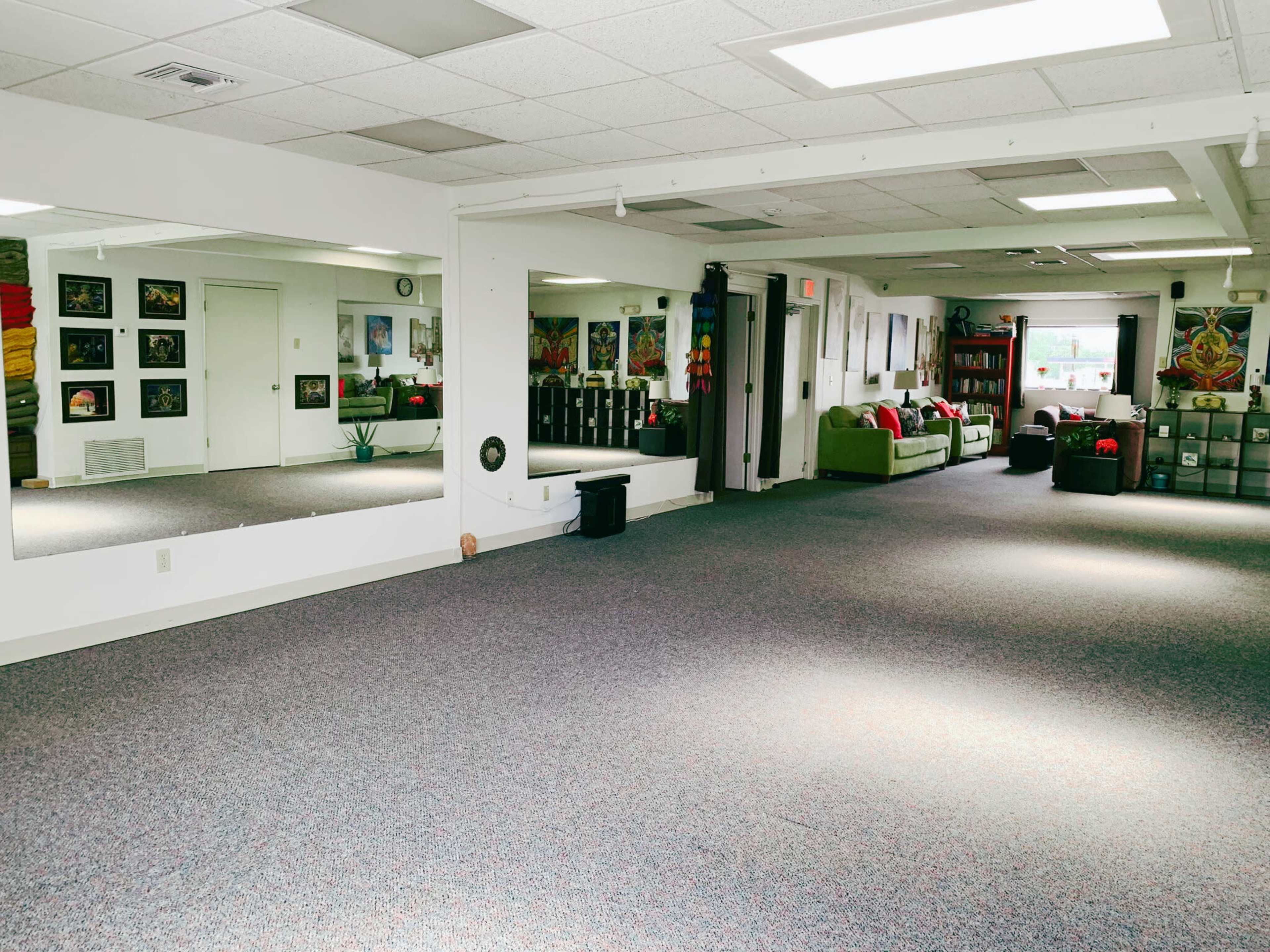 A spacious room with carpeted flooring, a mirror along one wall, and various artworks displayed, alongside seating areas and storage units in the background.