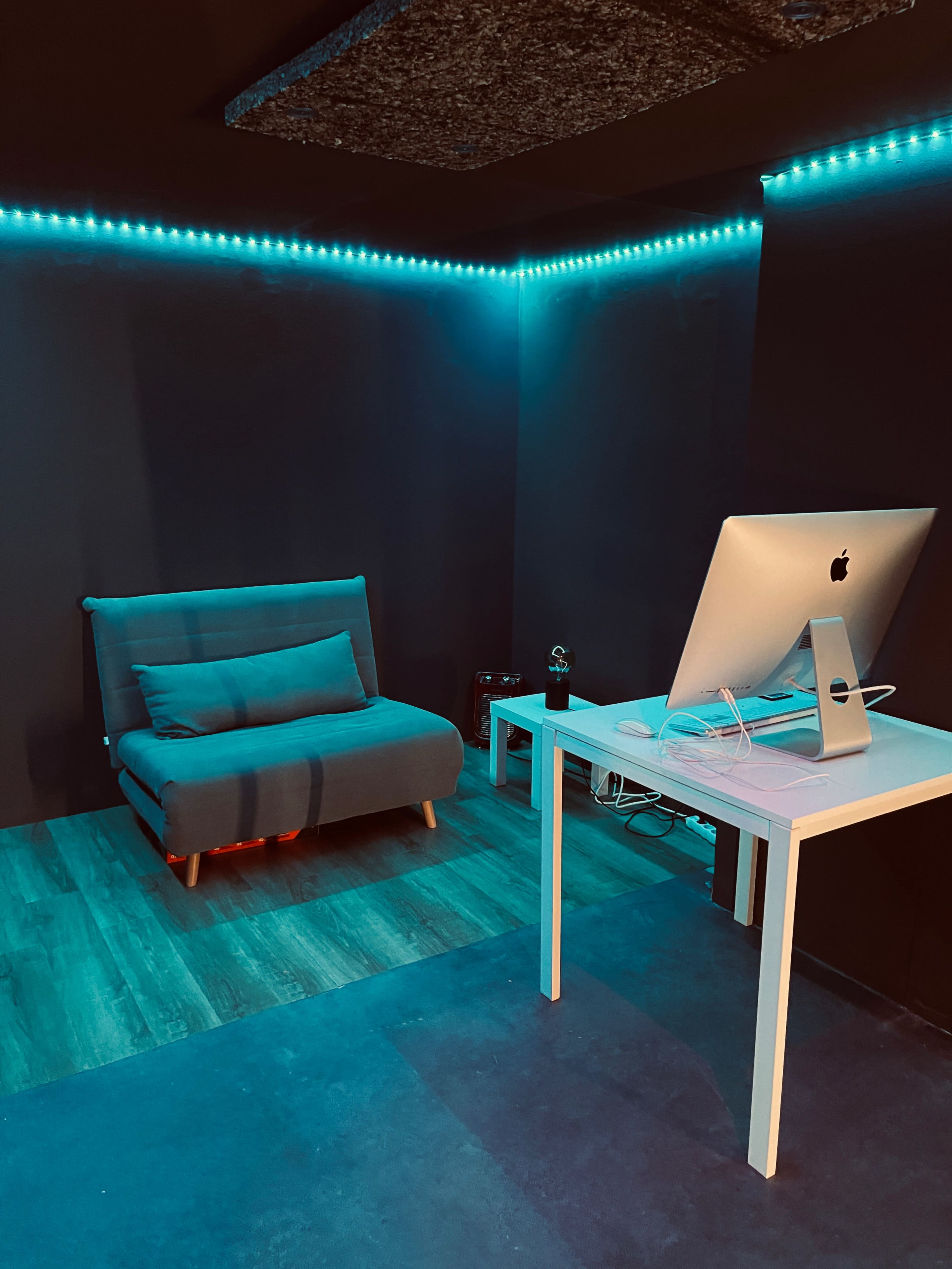 A small room with a blue-lit wall, featuring a gray sofa and a white desk with an iMac.