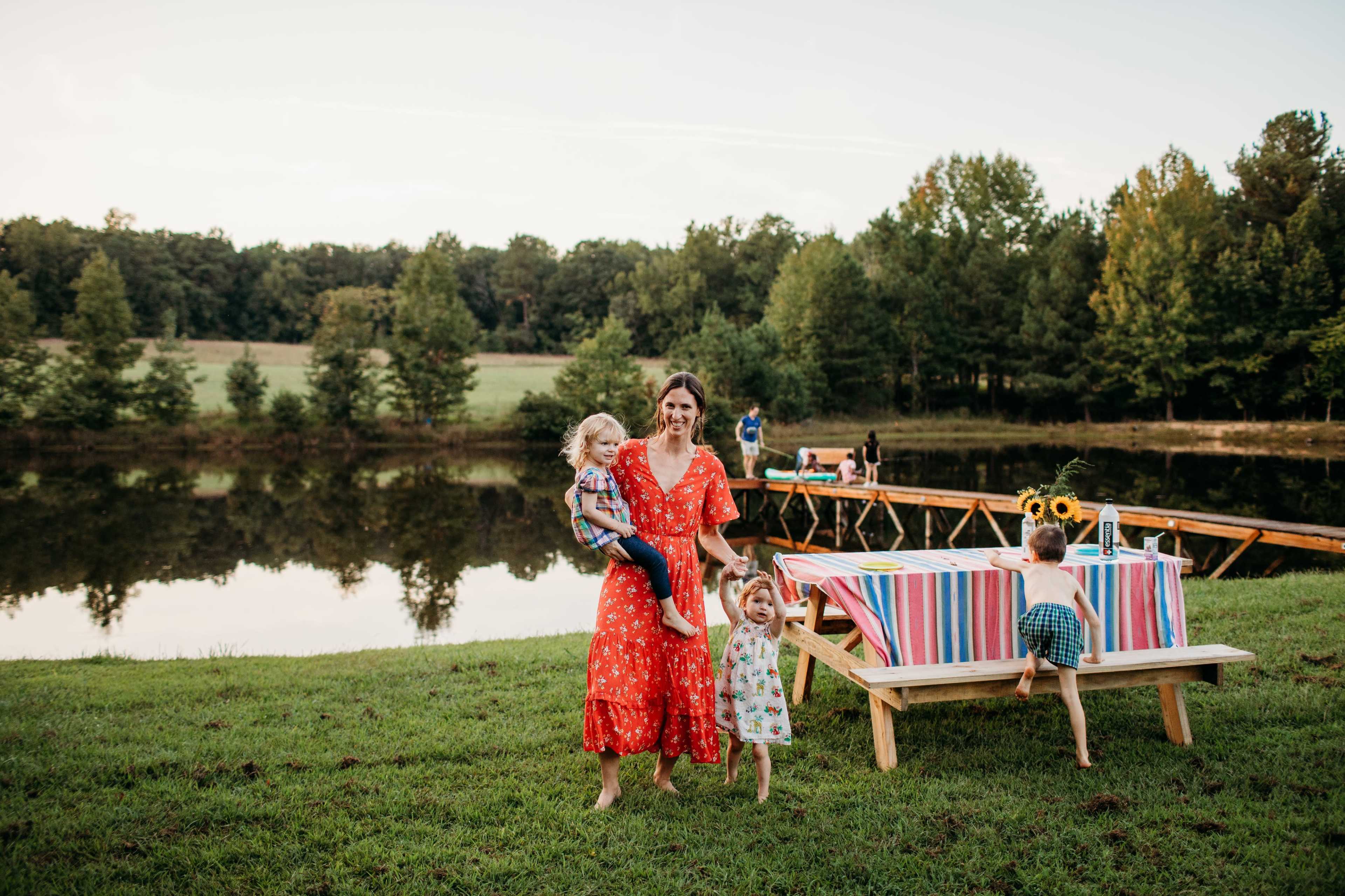 A woman in a red dress holds a child while another child plays nearby, with a picnic setup and people gathered by a pond in the background.