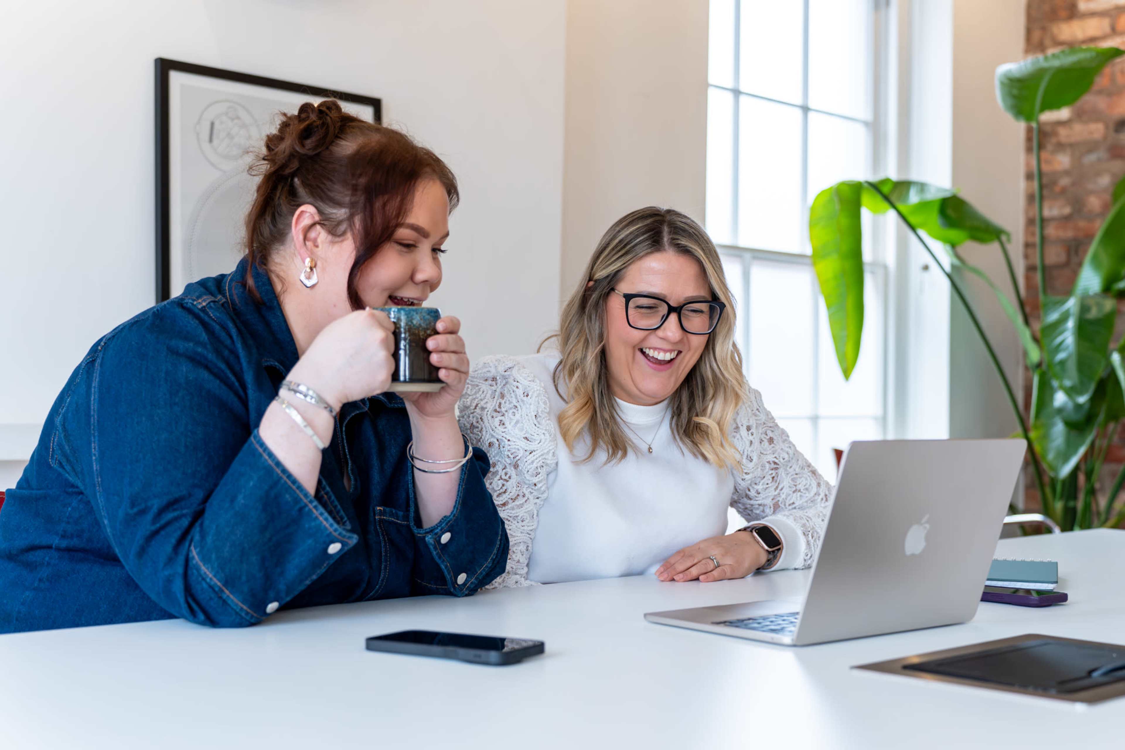 Two women sit at a table, smiling at a laptop while one holds a coffee cup.