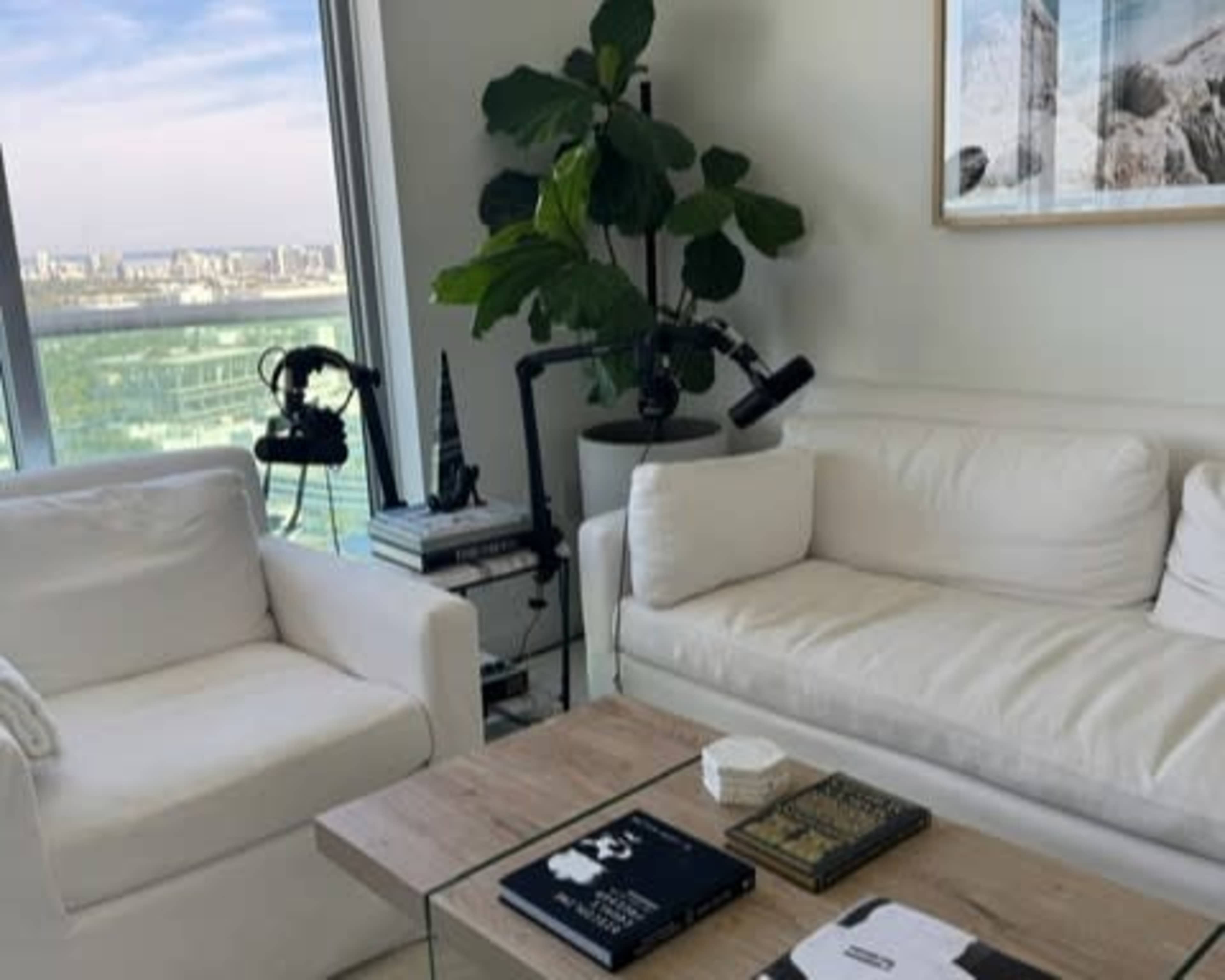 The image features a modern living room with two white sofas, a glass coffee table, and a large potted plant near a window that offers a city view.