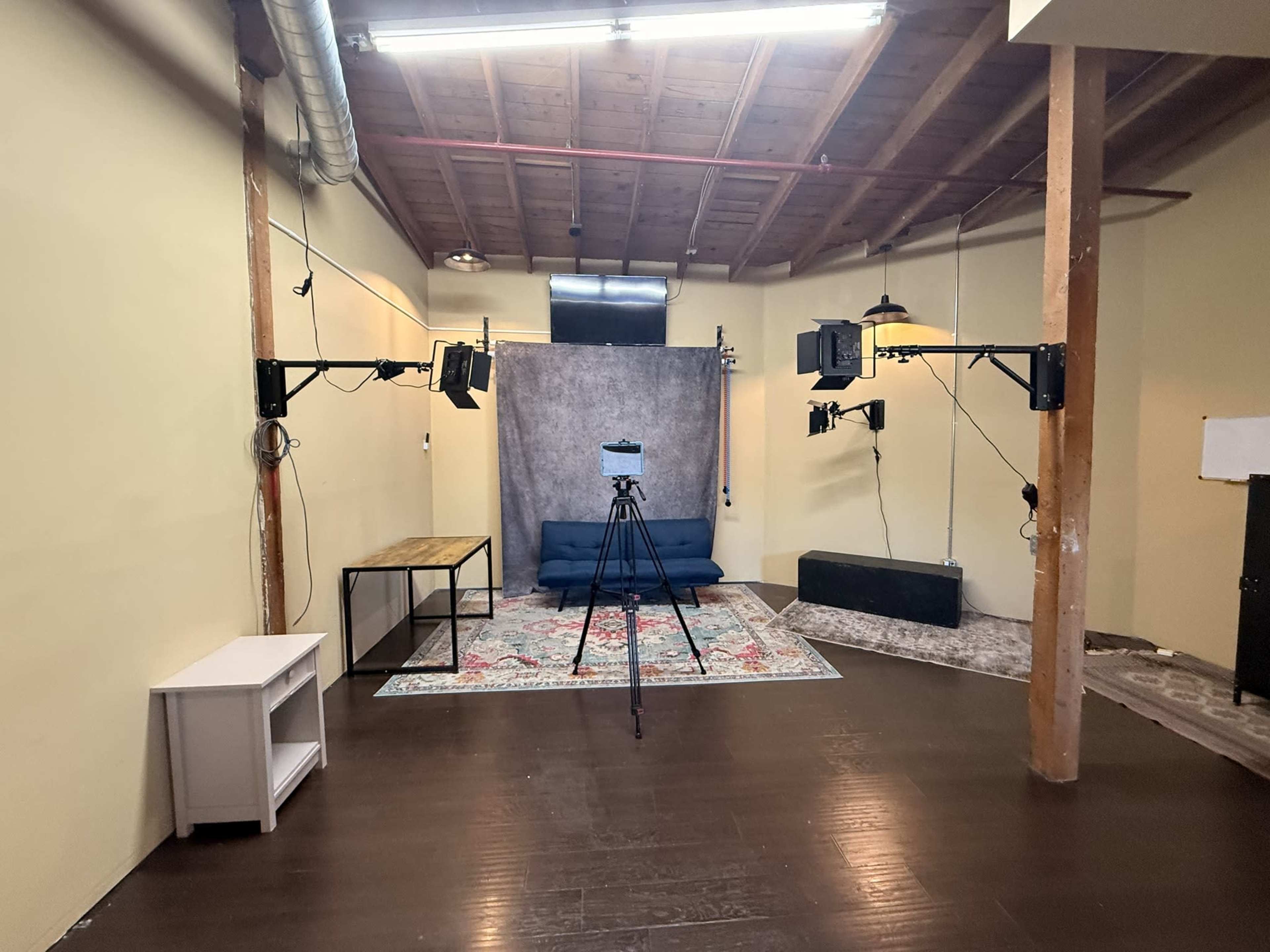 The image shows a spacious room with a wooden floor, equipped with a blue couch, a backdrop, several lights mounted on stands, and a tripod for a camera.