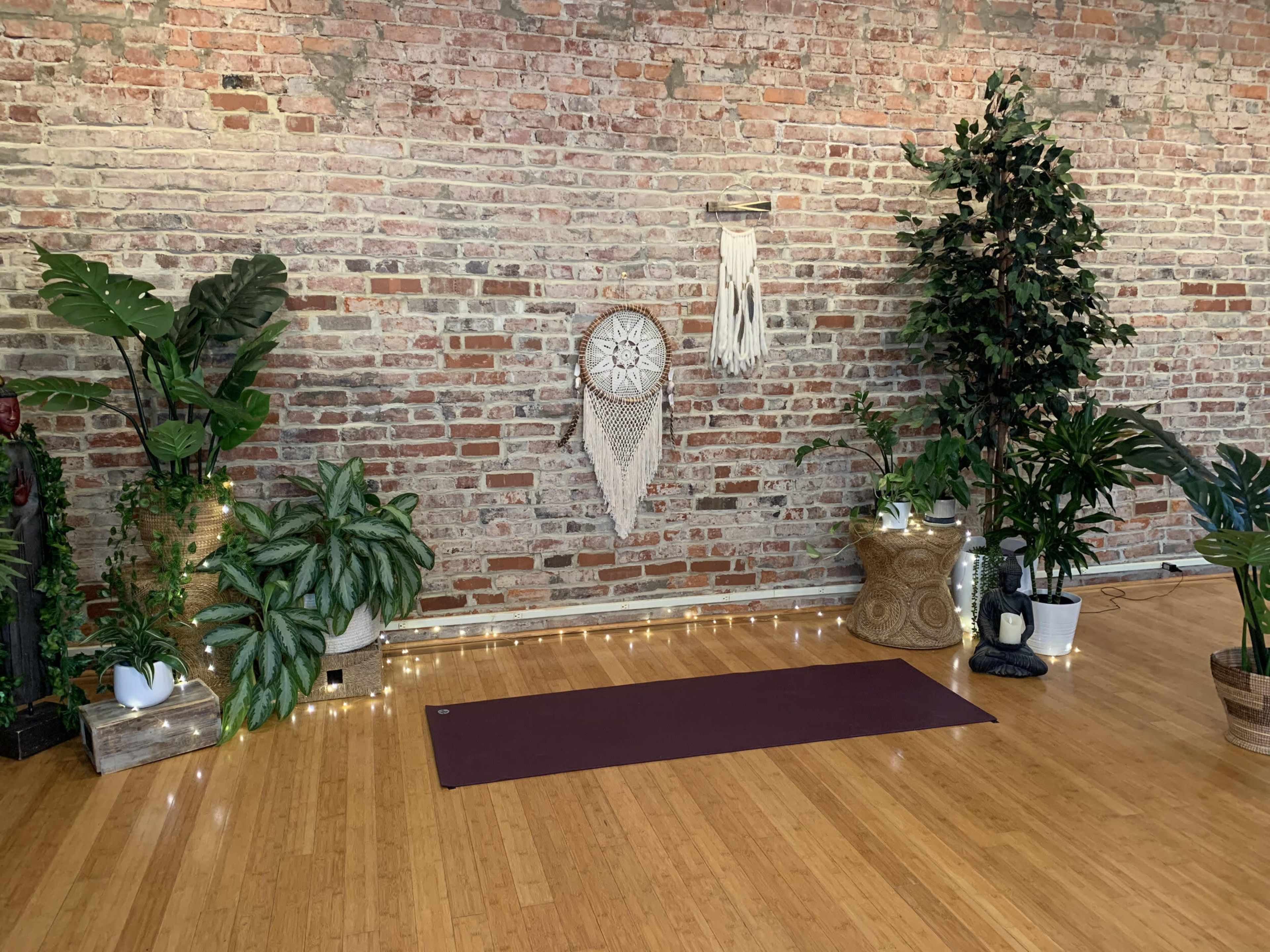 A yoga studio features a wooden floor with a maroon mat surrounded by various plants and decorative elements against a brick wall.