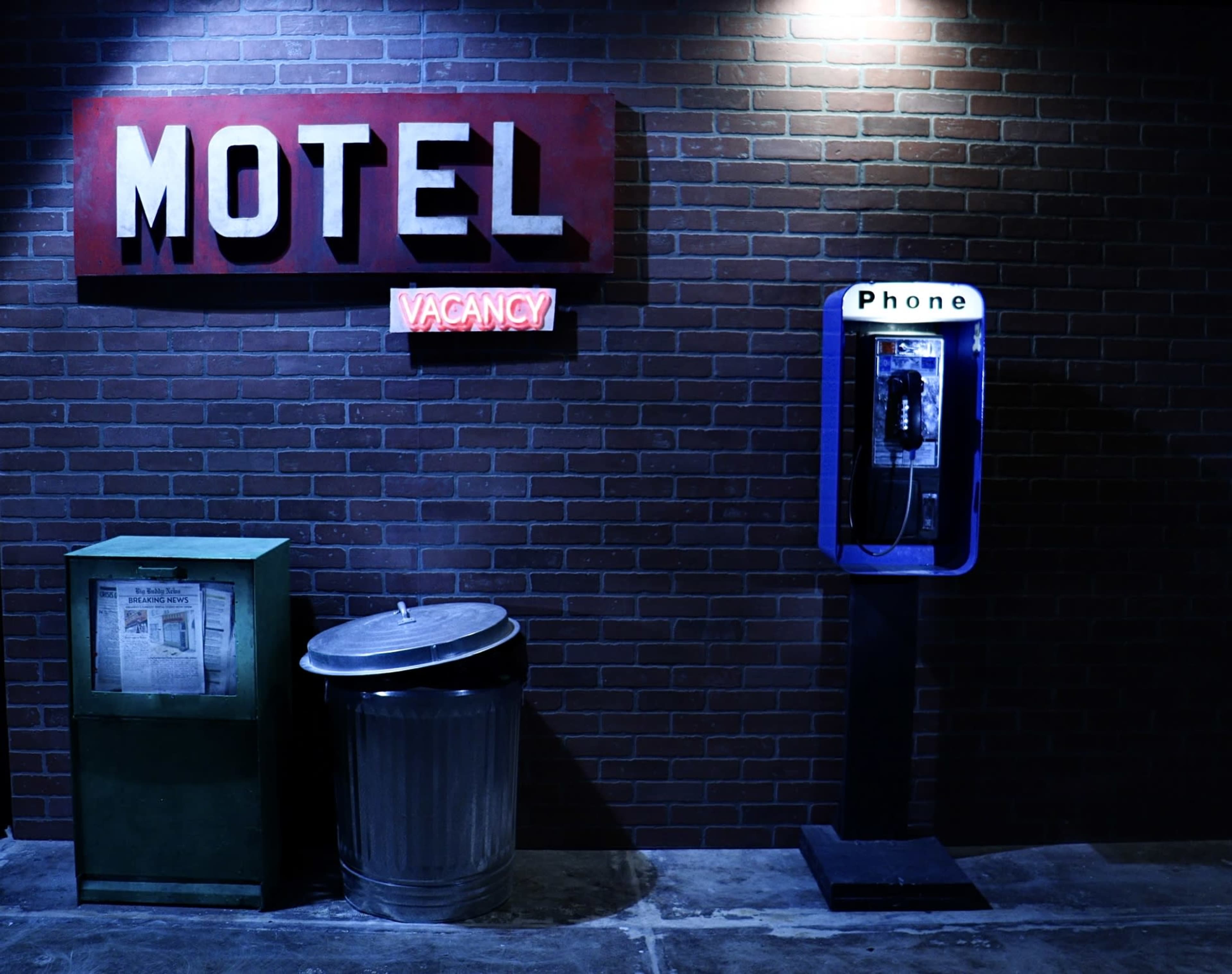 A vintage motel sign with a "Vacancy" light is positioned next to a payphone, a newspaper stand, and a trash can against a brick wall.