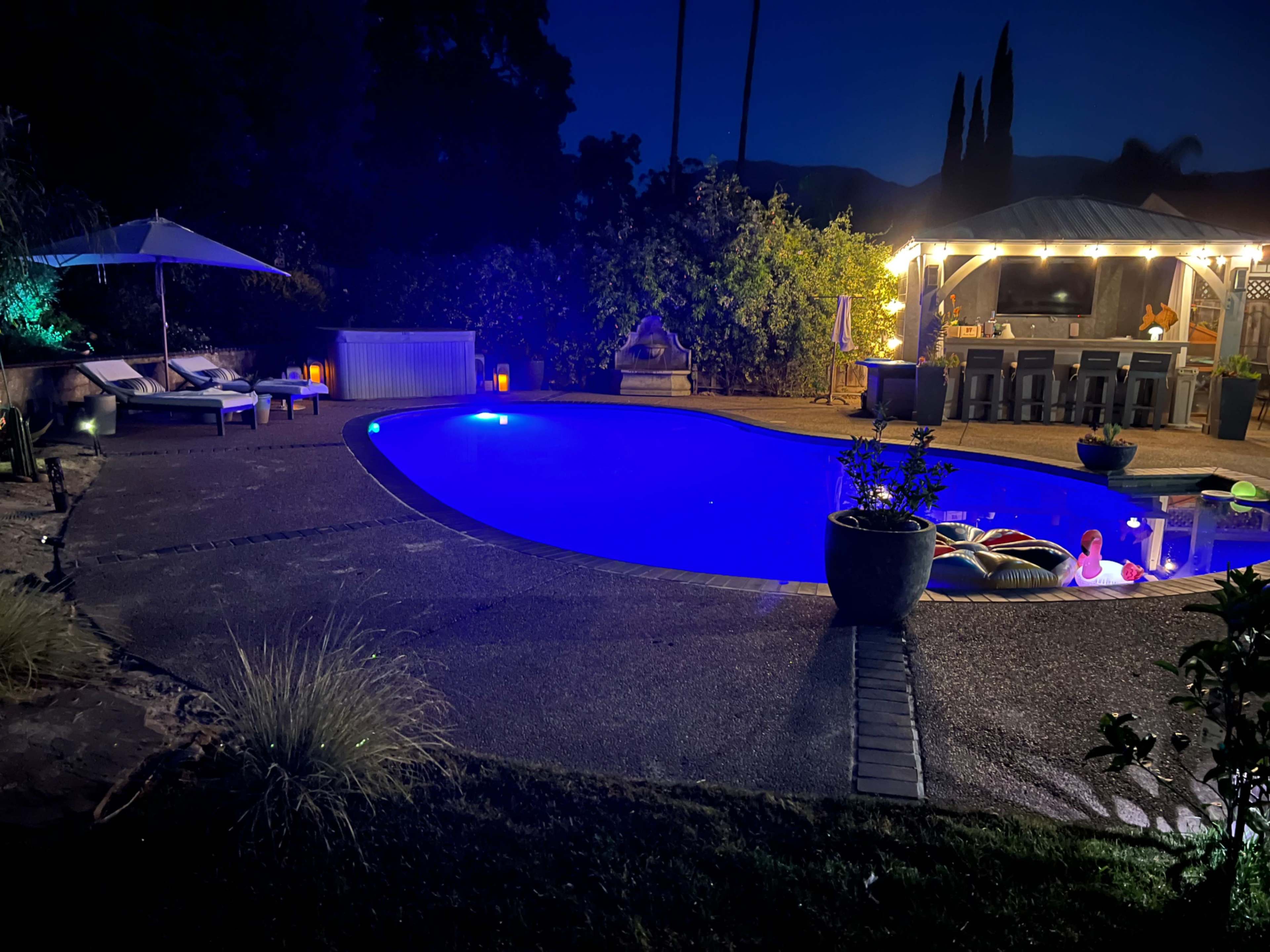 The image shows a illuminated swimming pool at night, surrounded by lounge chairs and a covered outdoor bar area with seating.