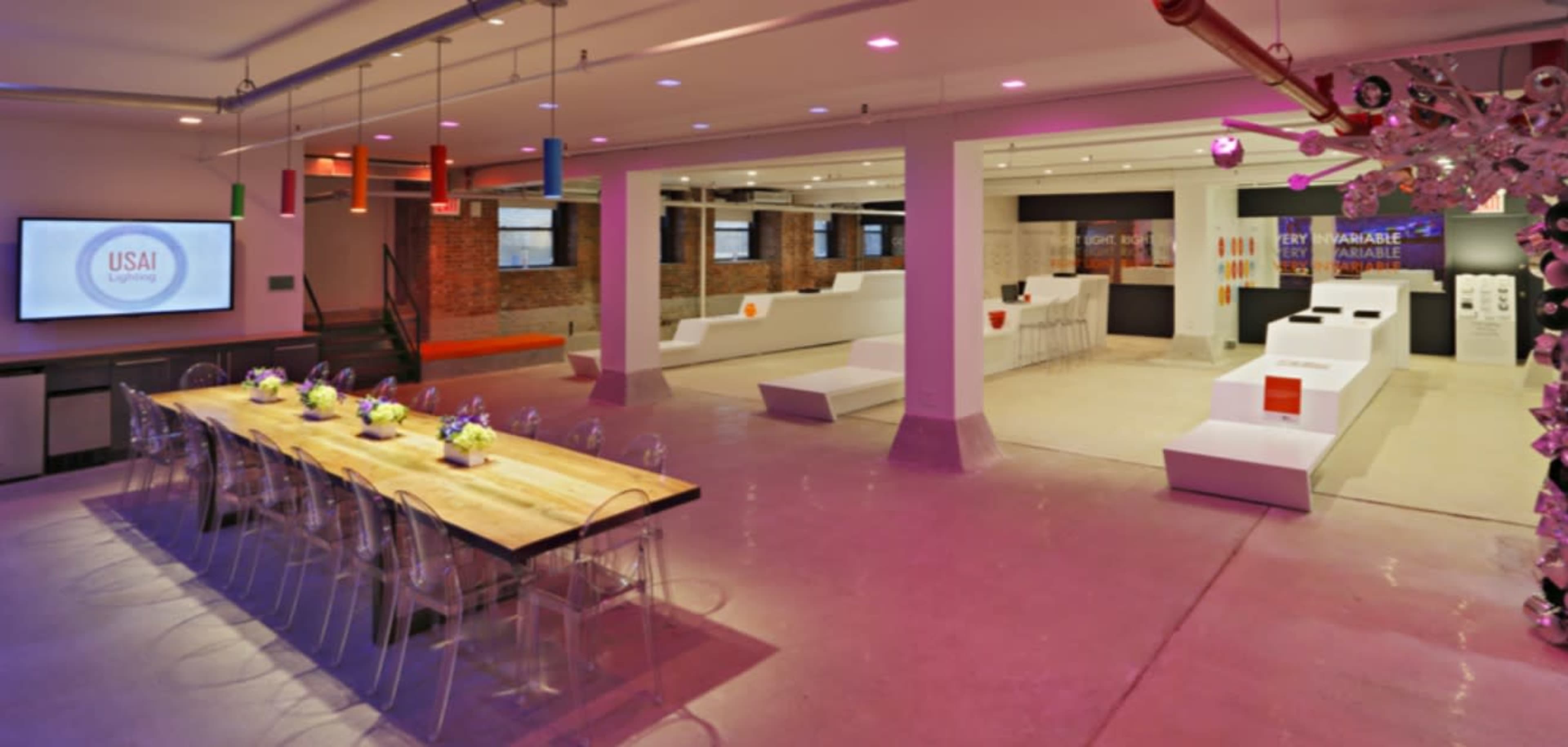 The image shows a modern event space featuring a long wooden dining table with transparent chairs and an open area with minimalist white displays and colorful hanging lights.