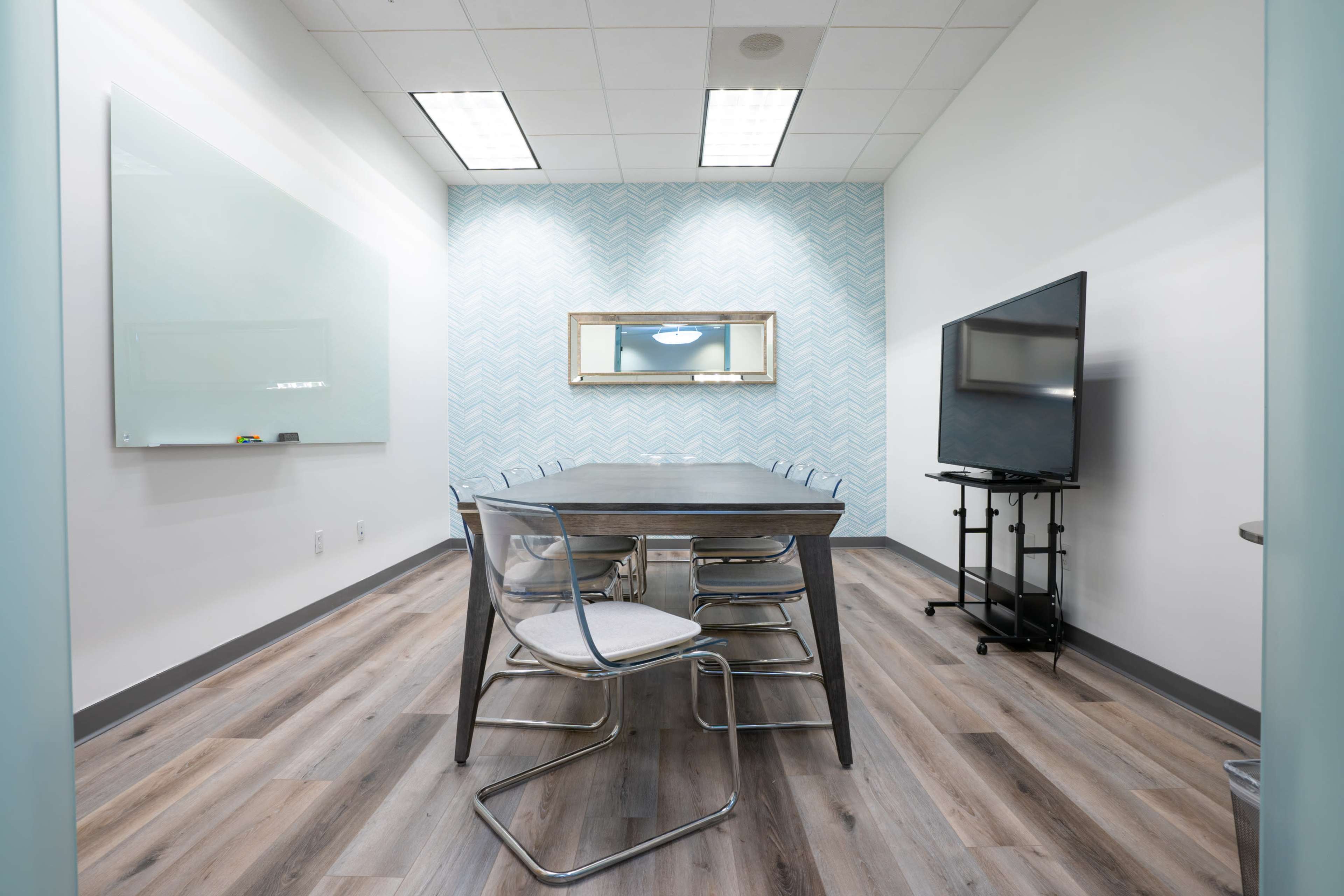 A small meeting room features a rectangular table surrounded by transparent chairs, a wall-mounted television, and light blue patterned wallpaper.