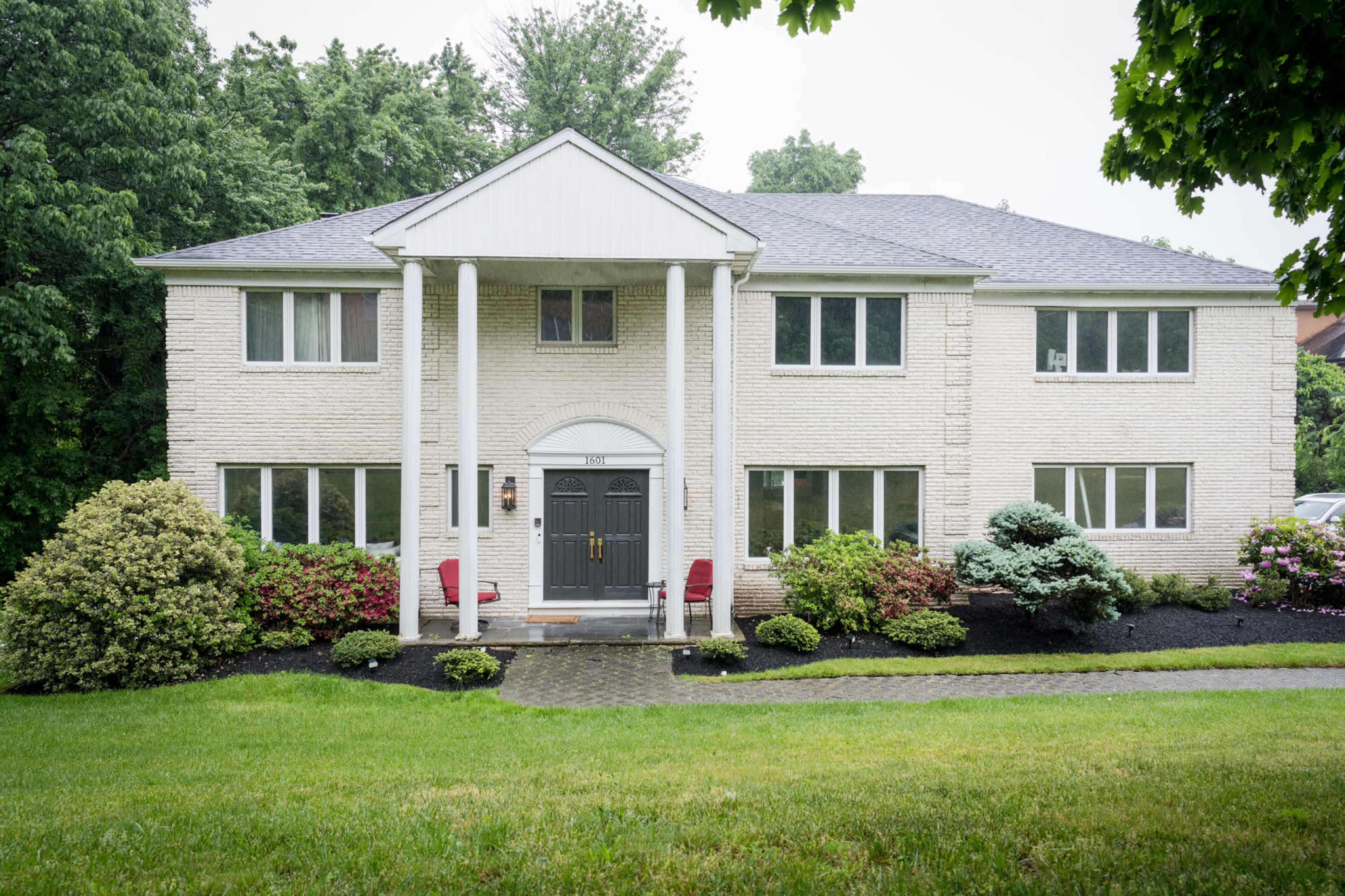 The image shows a two-story brick house with a central entrance, flanked by red chairs and surrounded by landscaped shrubs and grass.