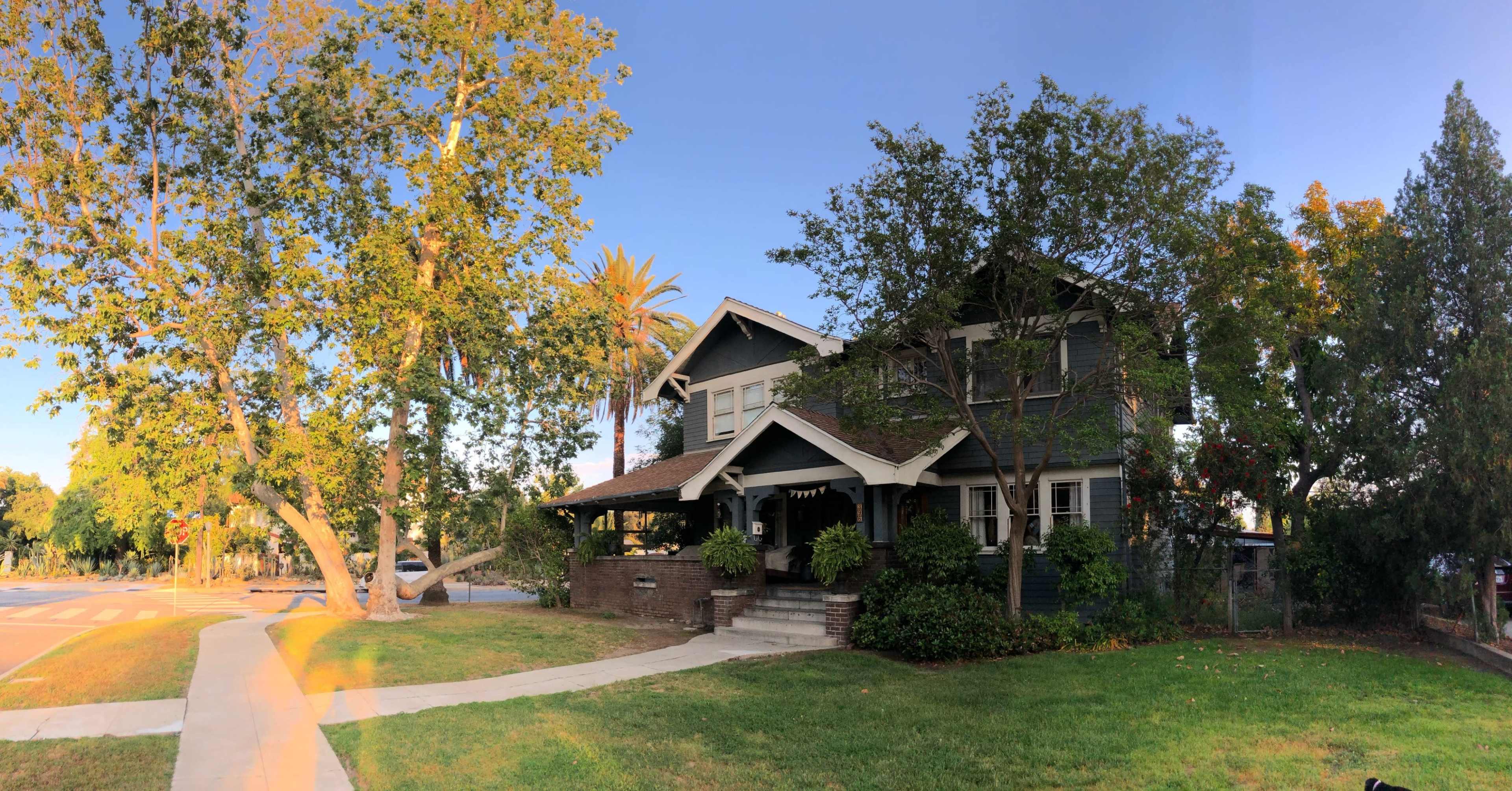 A two-story house with a dark exterior, large windows, and a wide porch is surrounded by trees and grassy landscaping on a corner lot.