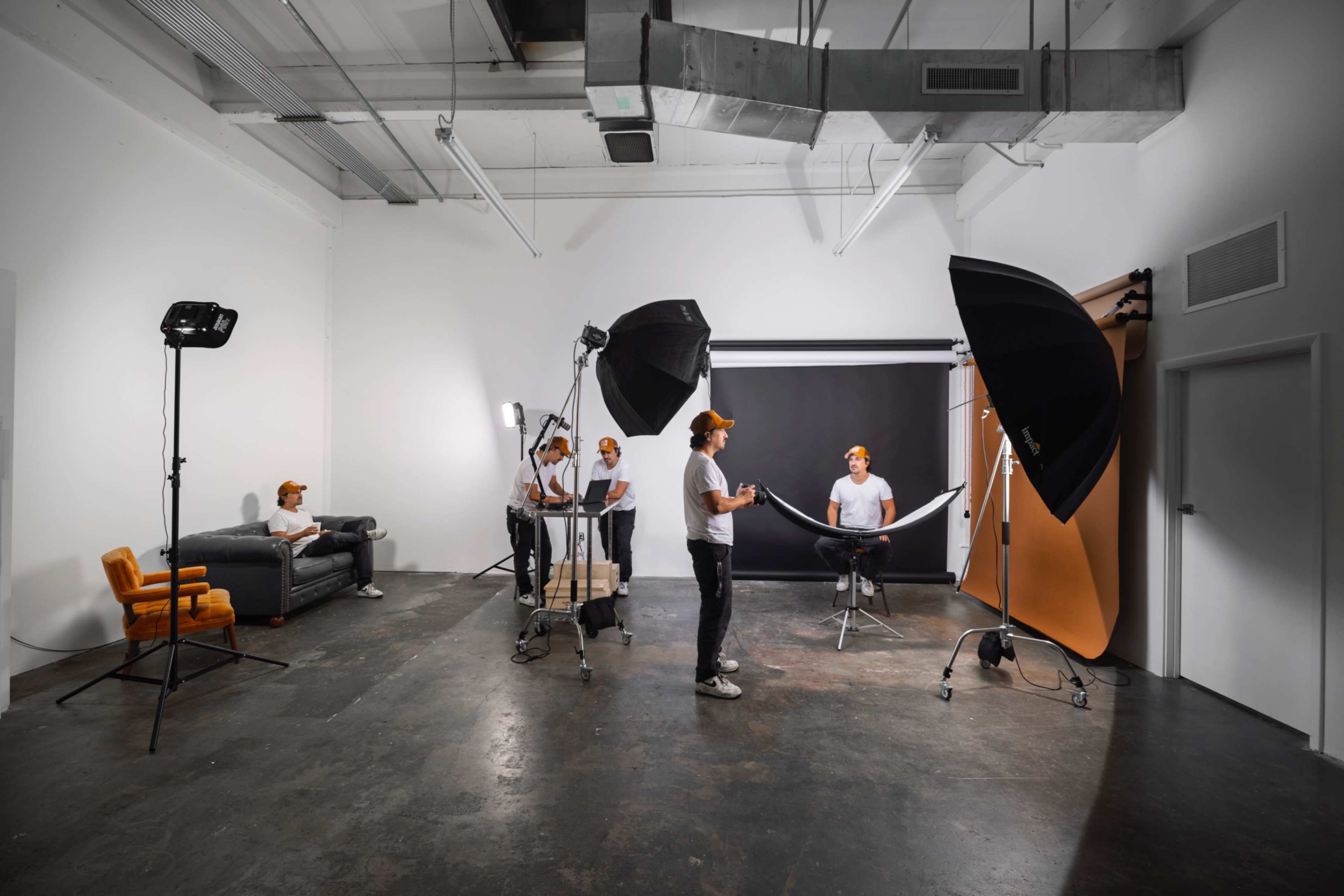 A photography studio features multiple lighting setups and a model posing in front of a backdrop, with assistants adjusting equipment.