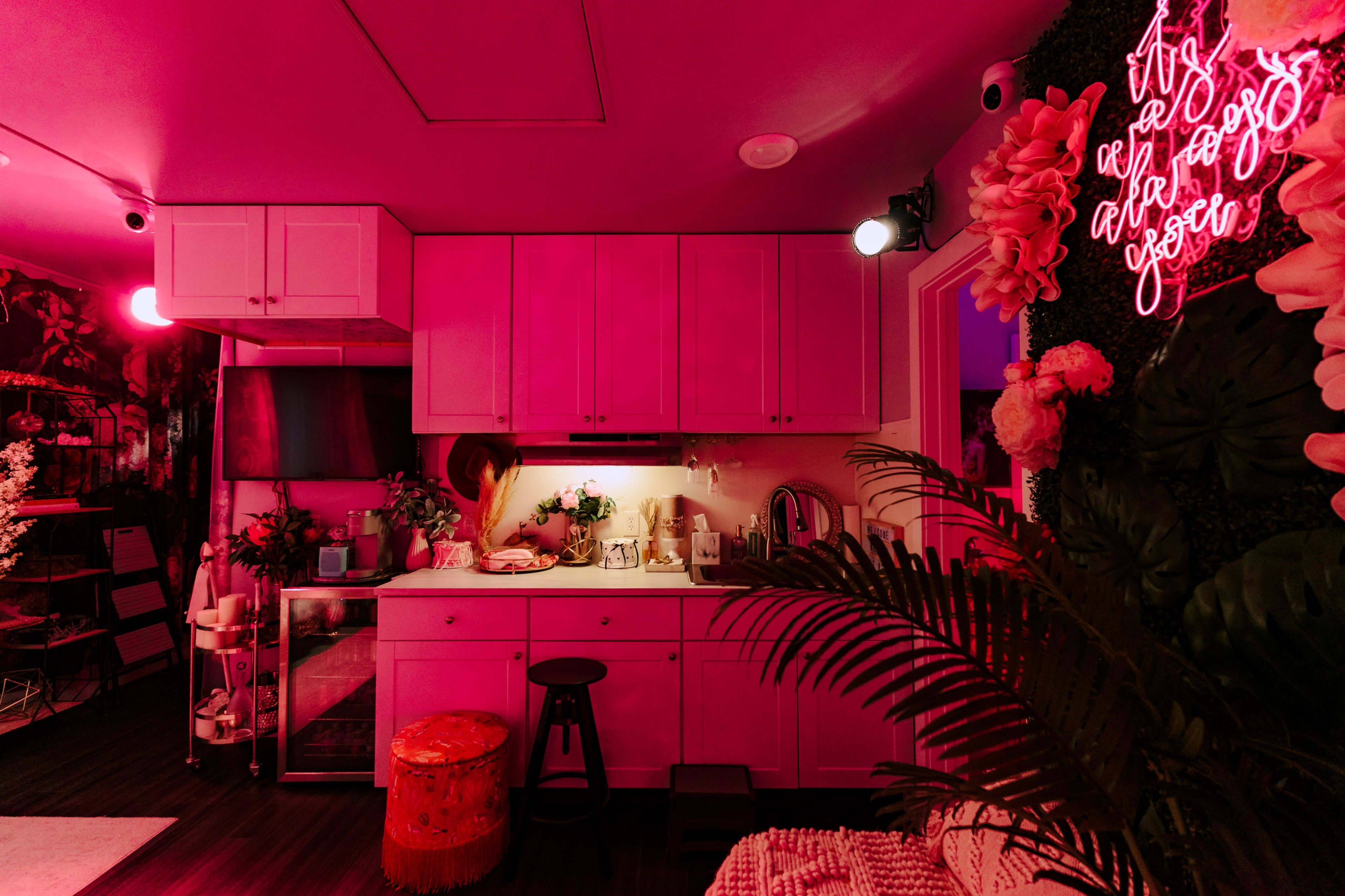 A modern kitchen with pink lighting features a woman preparing food, surrounded by decorative plants and a neon sign.