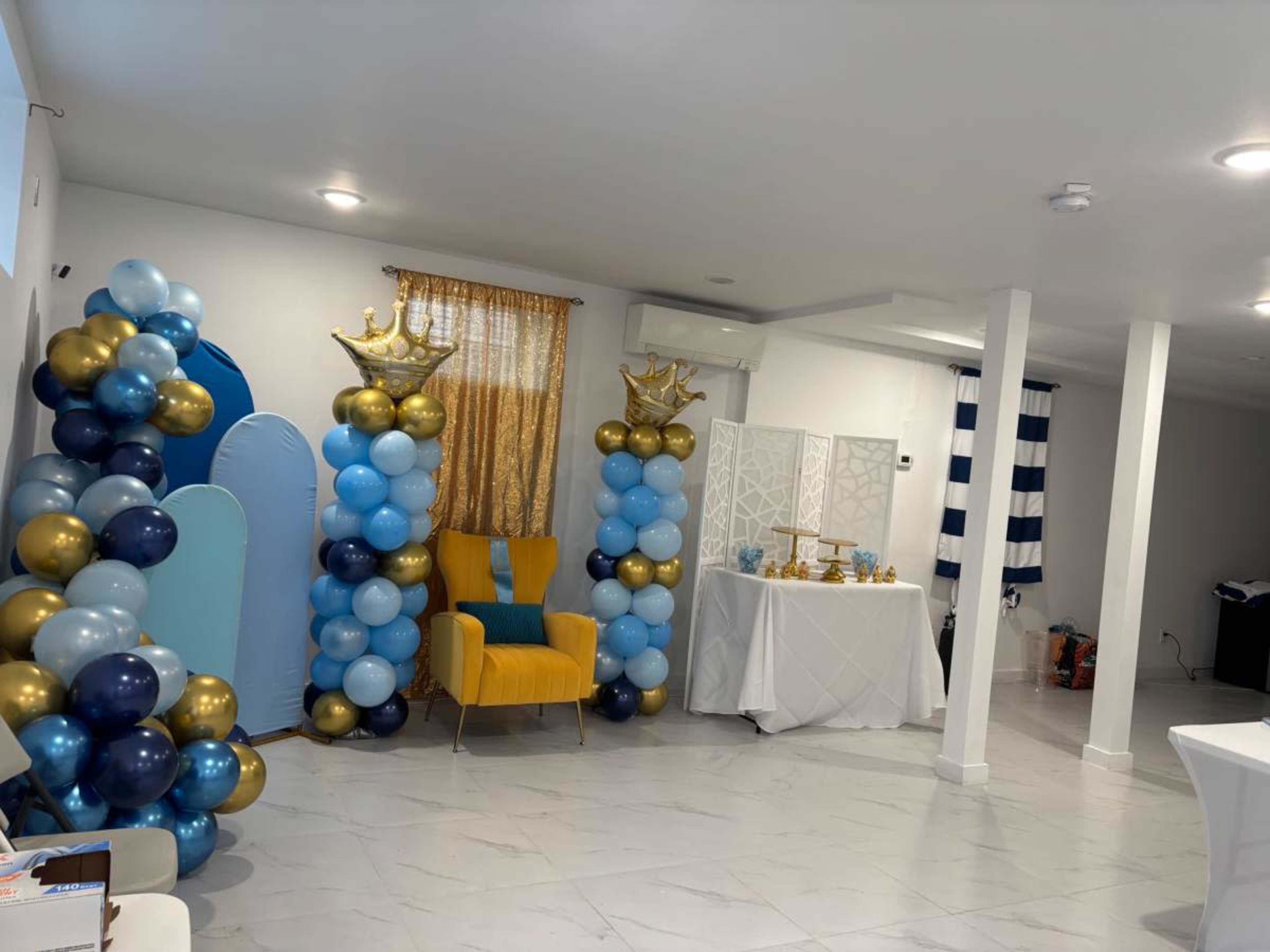 The scene features a decorated room with blue, gold, and turquoise balloons arranged in columns, a yellow chair, and a white table with festive items.
