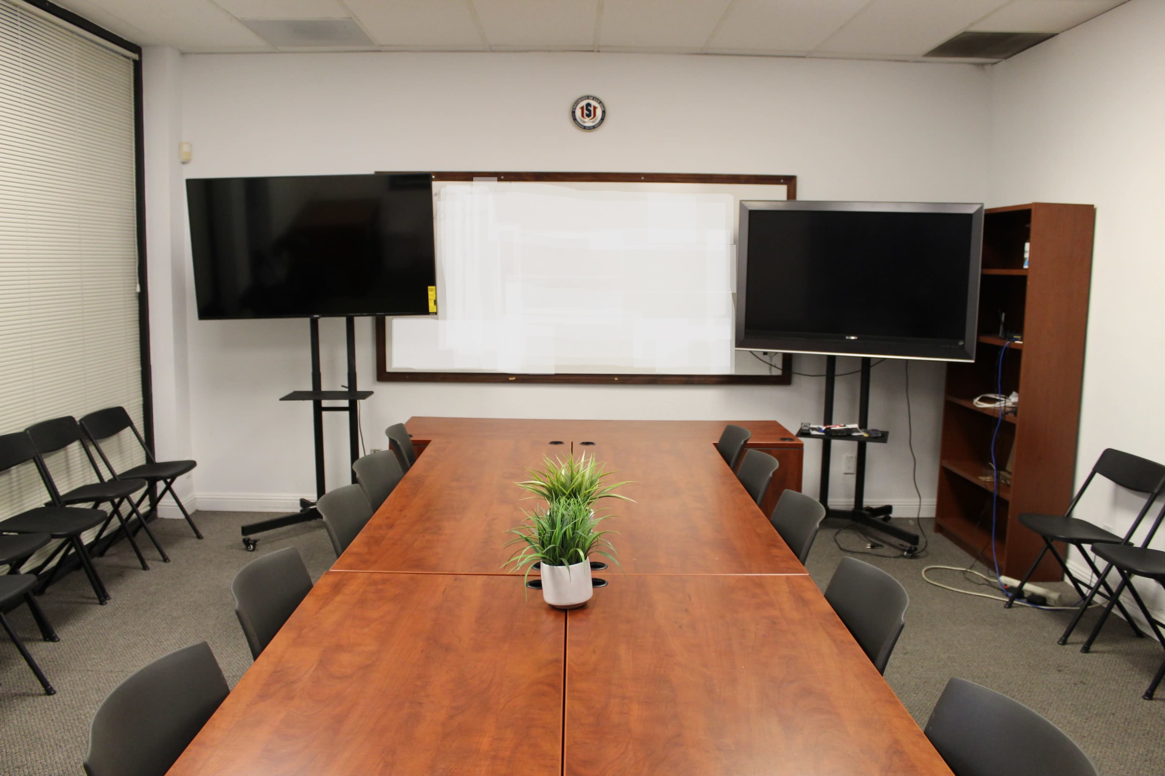 A conference room features a long wooden table, two large monitors on stands, a whiteboard, and black folding chairs arranged neatly around the table.