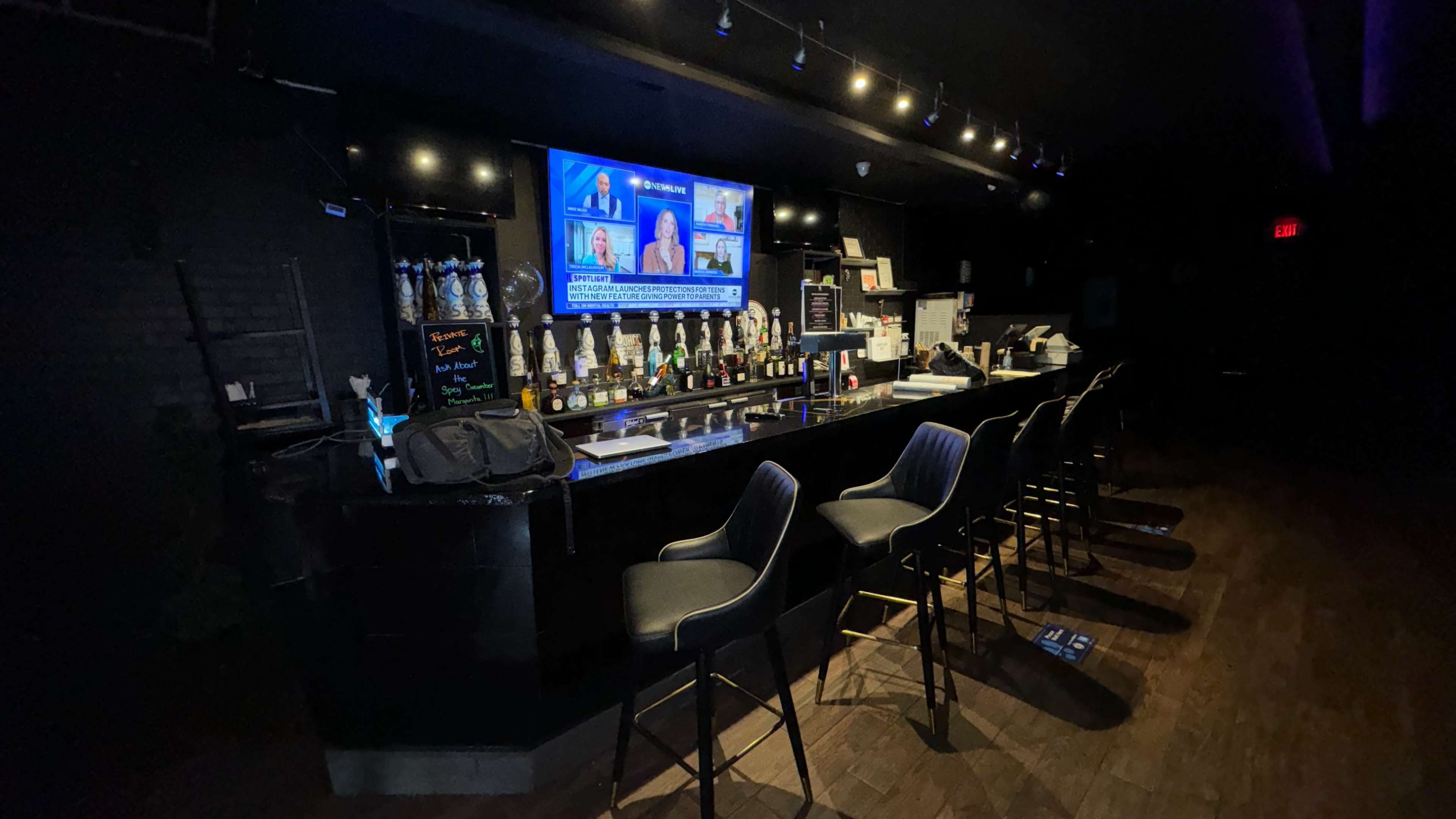 A dimly lit bar features a long counter with several empty black stools and a large screen displaying news broadcasts.