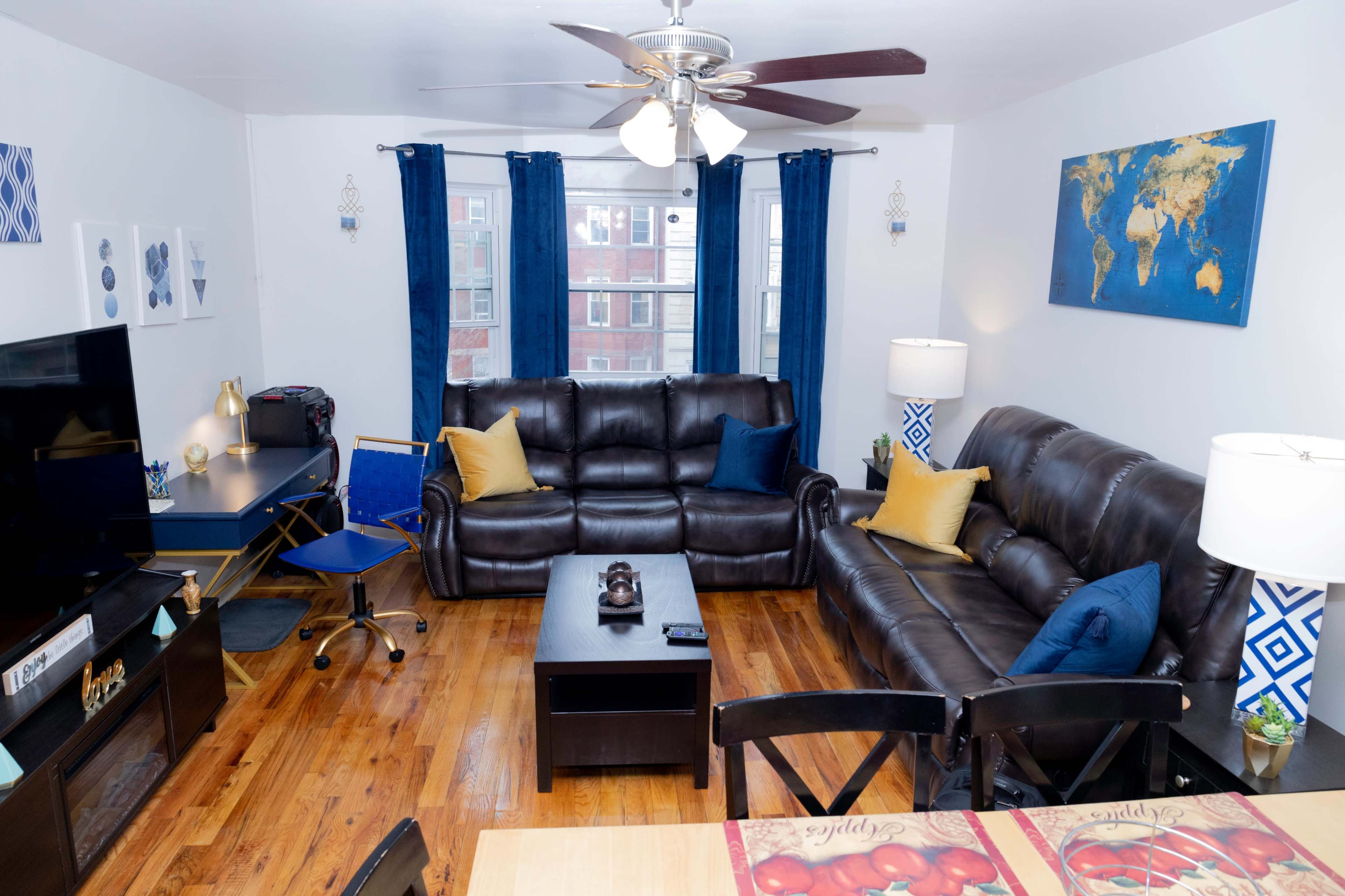 A living room features two dark leather sofas, a coffee table, a TV stand, and a desk, with blue curtains framing the windows and a world map on the wall.