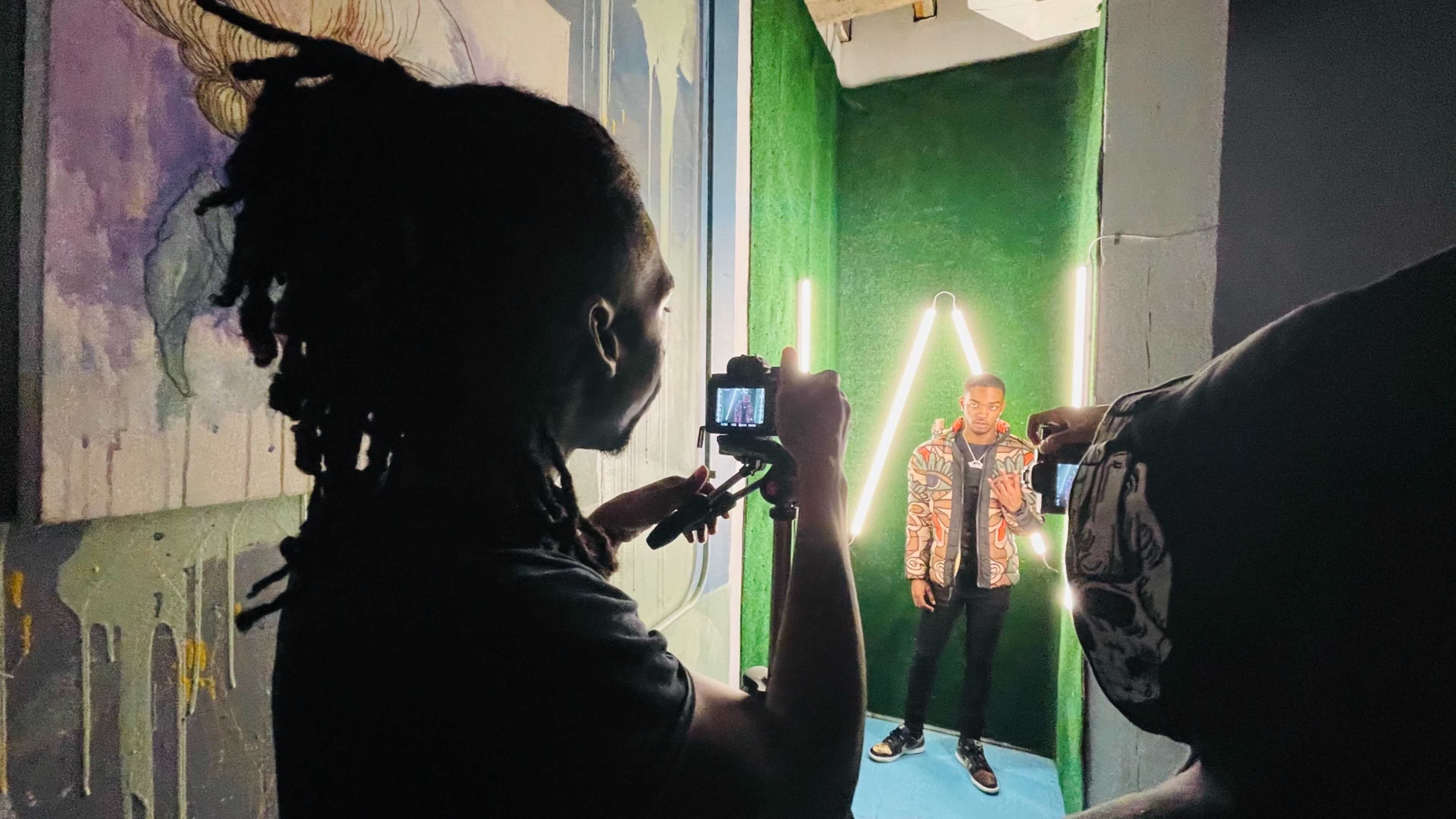 A person is taking photos of another person standing in a brightly lit corner with green walls and neon lights.