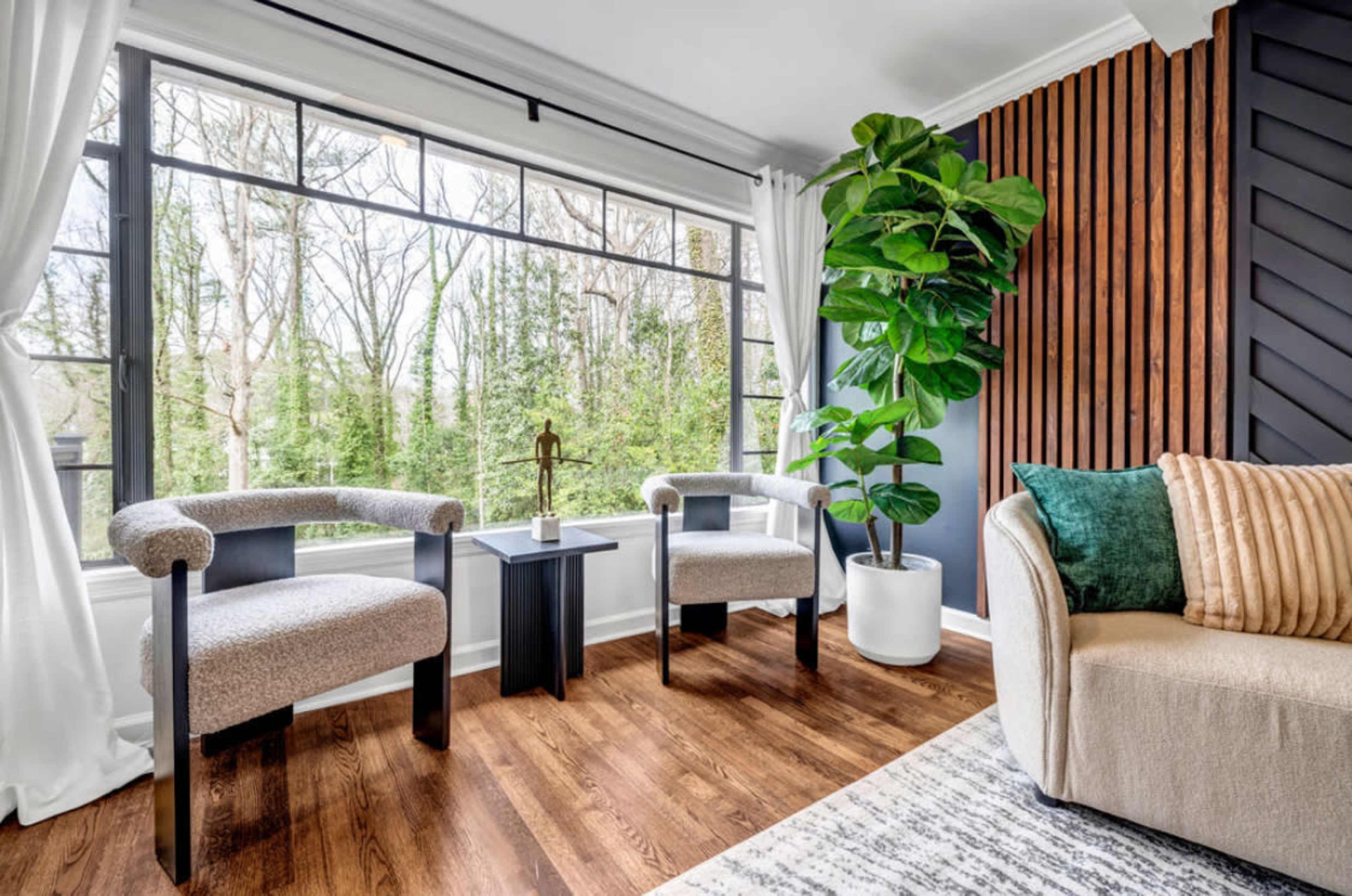 The image shows a modern living space with two upholstered chairs, a small side table, and a large potted plant near a large window overlooking a wooded area.