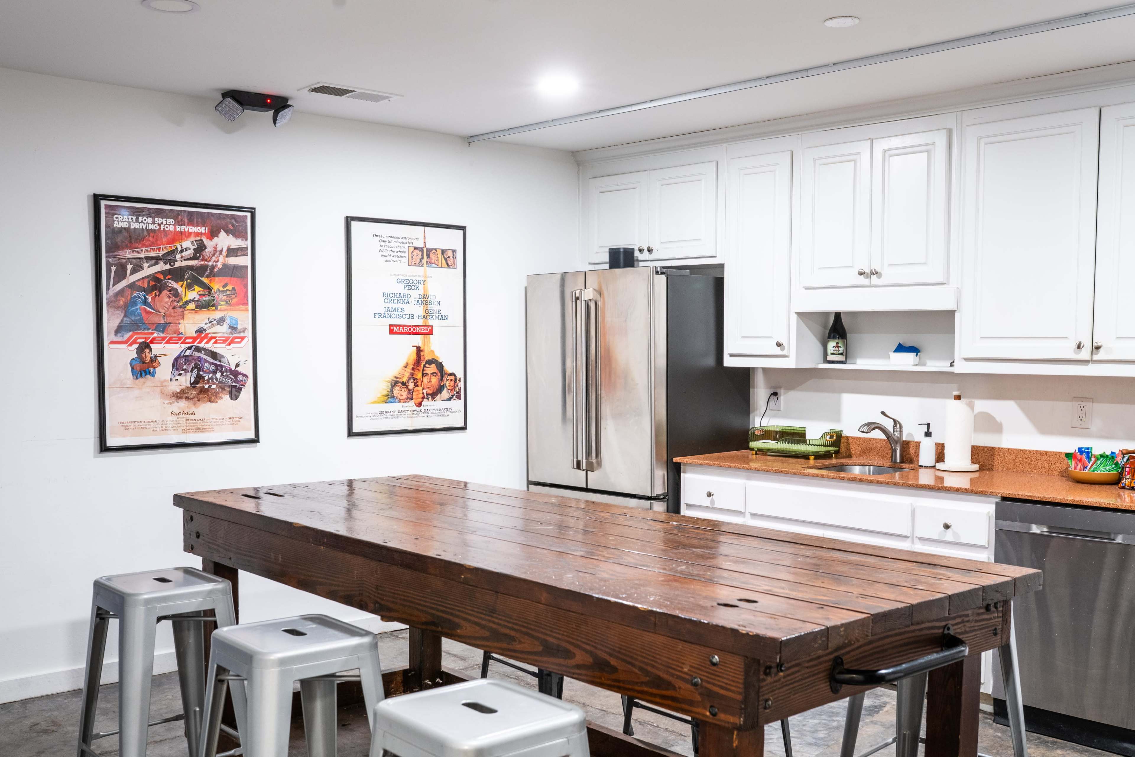 A kitchen area with a wooden table and metal stools, featuring framed movie posters on the wall and a stainless steel refrigerator.