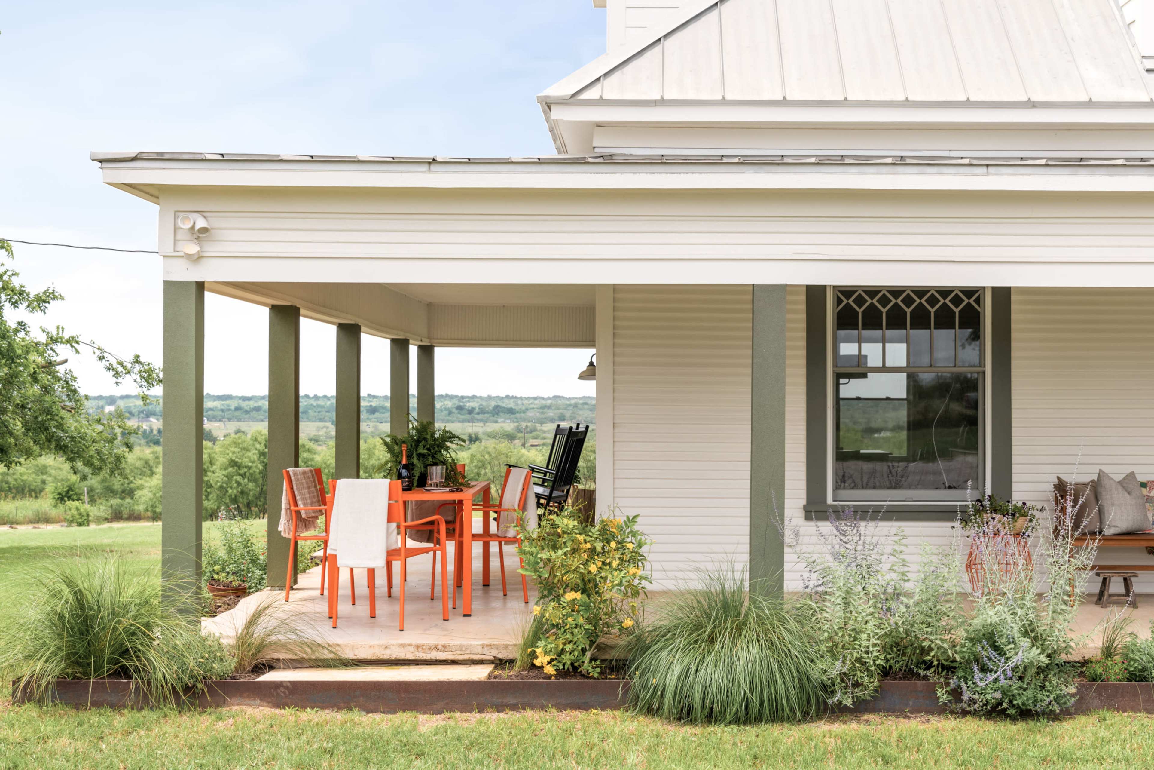 A covered porch with orange chairs and a dining table overlooks a grassy field and distant hills.