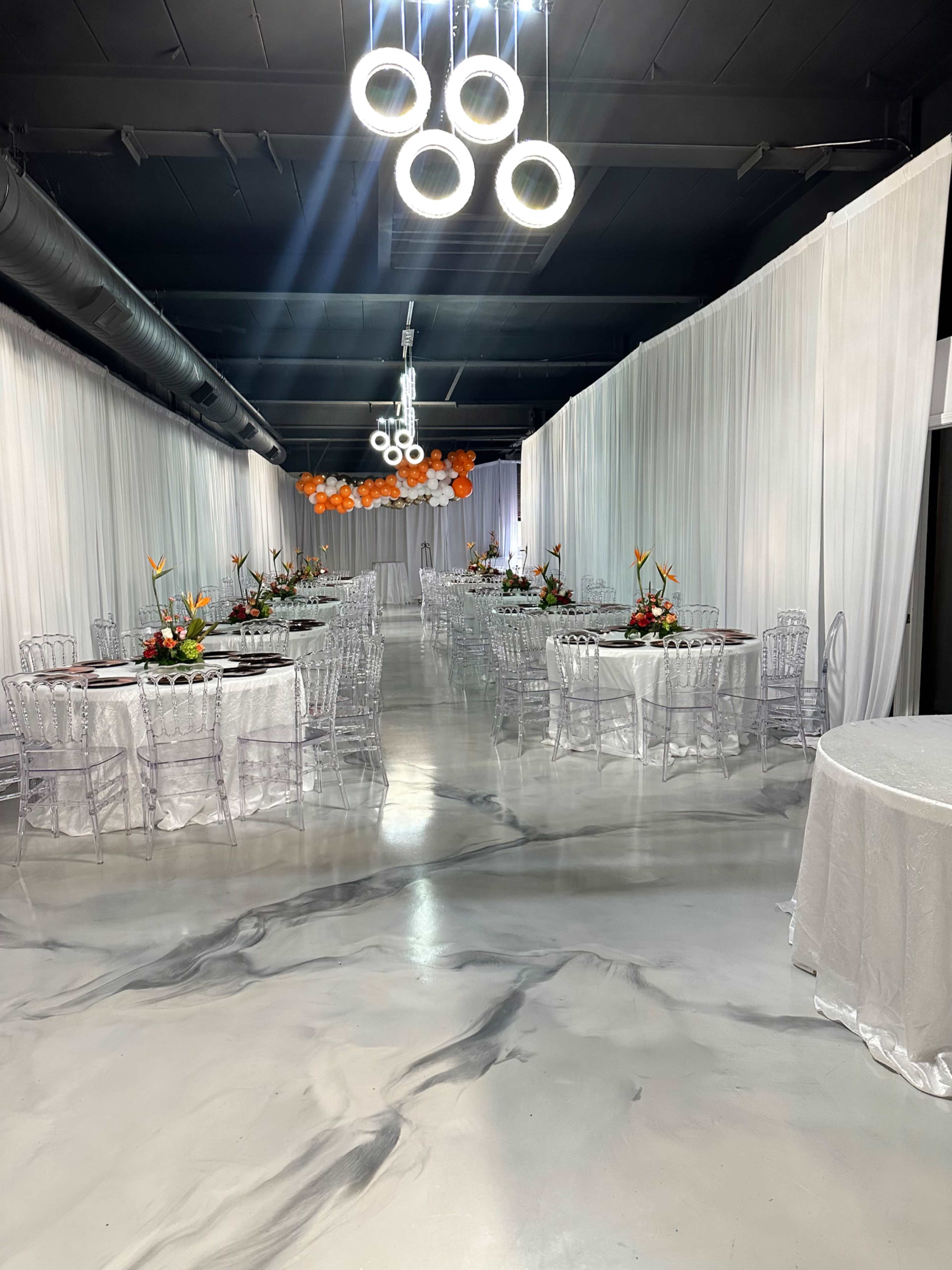 The image shows a spacious event hall with white draped walls, circular chandeliers, and decorated tables arranged in two rows.