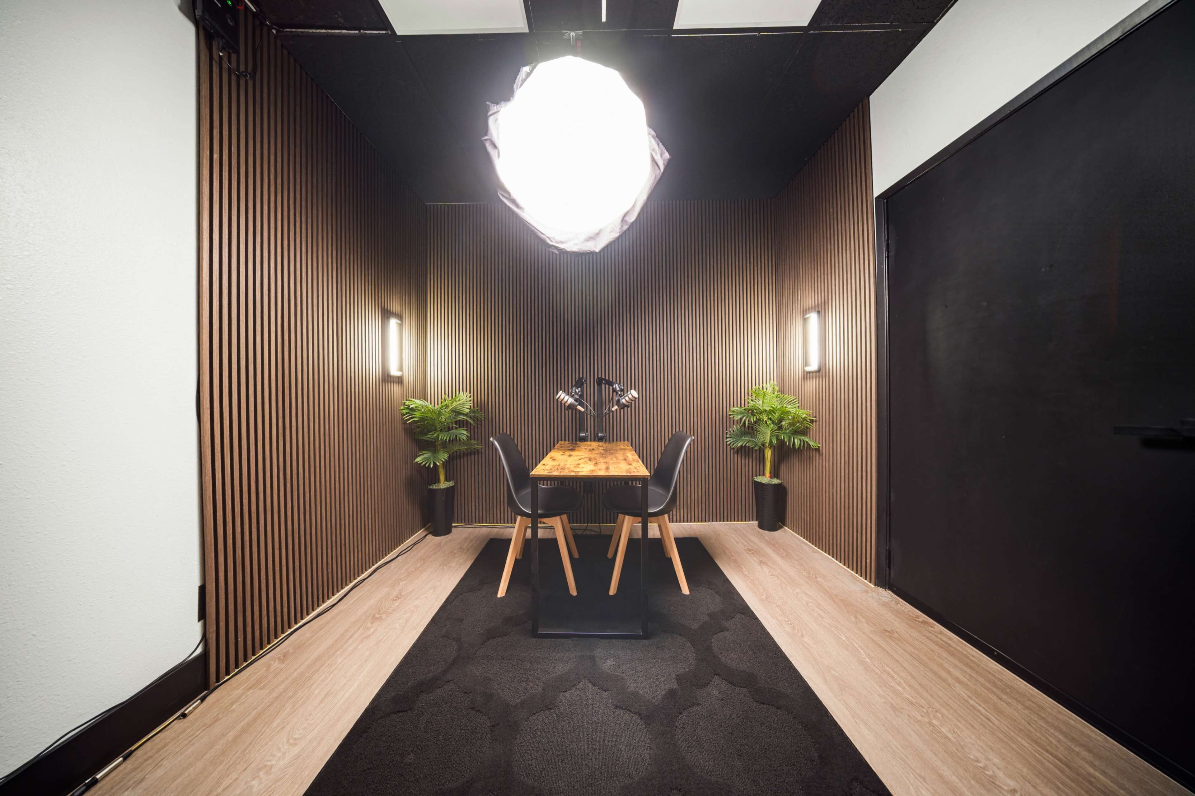 The image shows a modern, minimalistic room with a wooden table and chairs, flanked by potted plants, set against a backdrop of dark wooden paneling and a black wall.