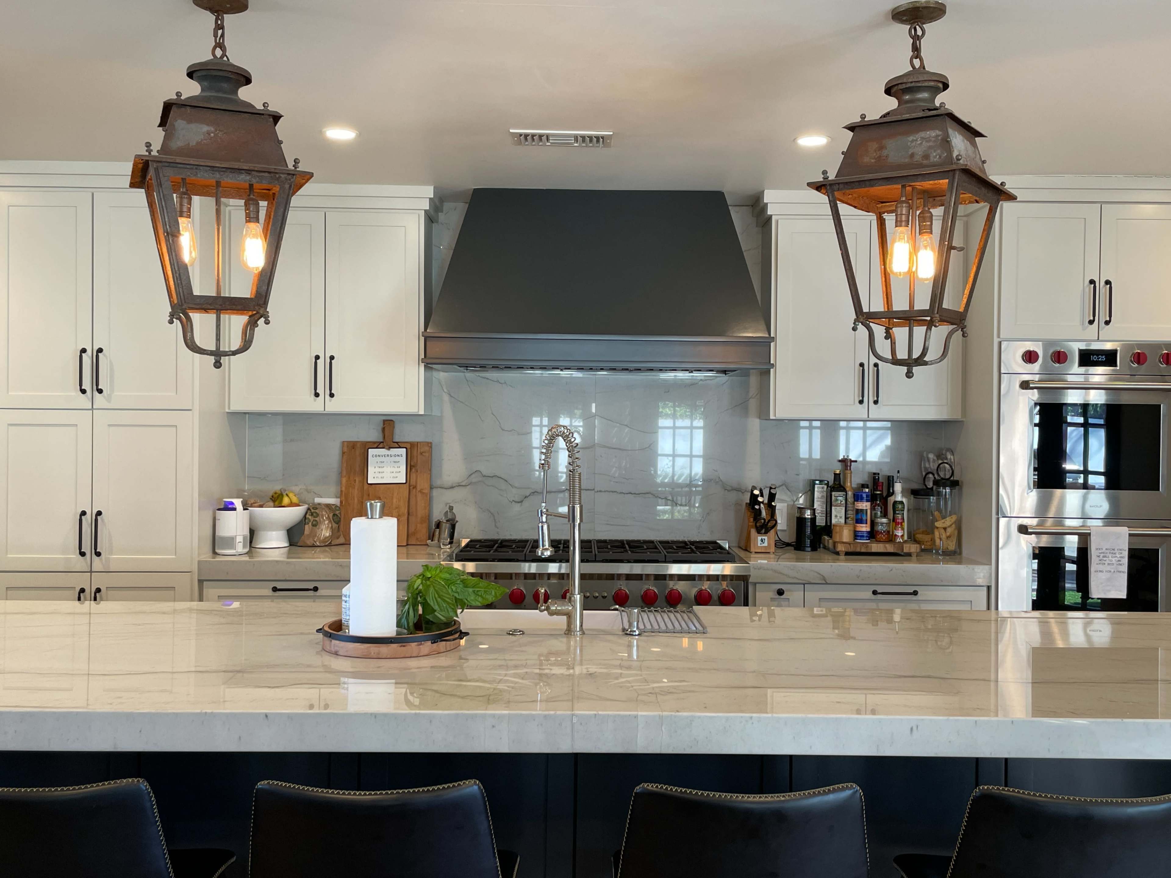 A modern kitchen featuring a large marble island, vintage-style pendant lights, and a stainless steel range hood above a gas stove.
