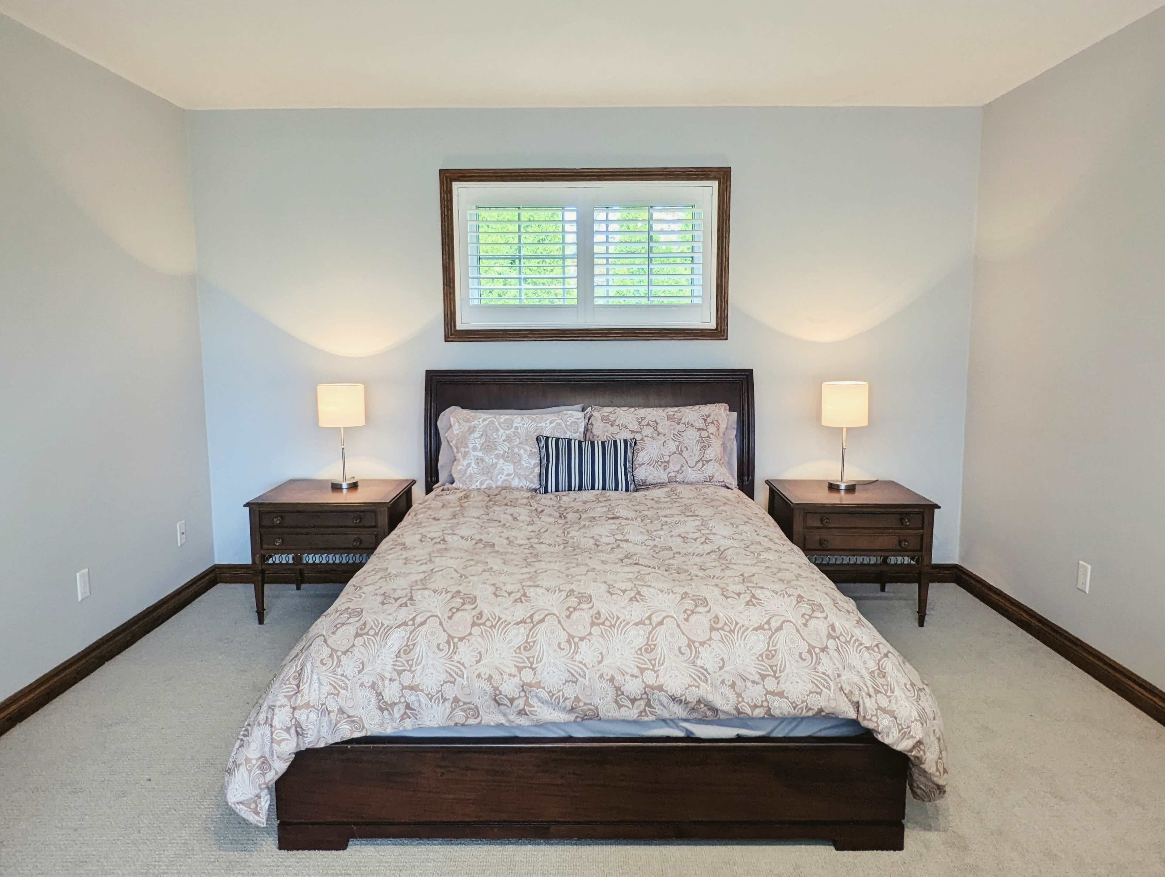 A neatly arranged bedroom features a large bed with a patterned comforter, flanked by two bedside tables with lamps, and a window above providing natural light.