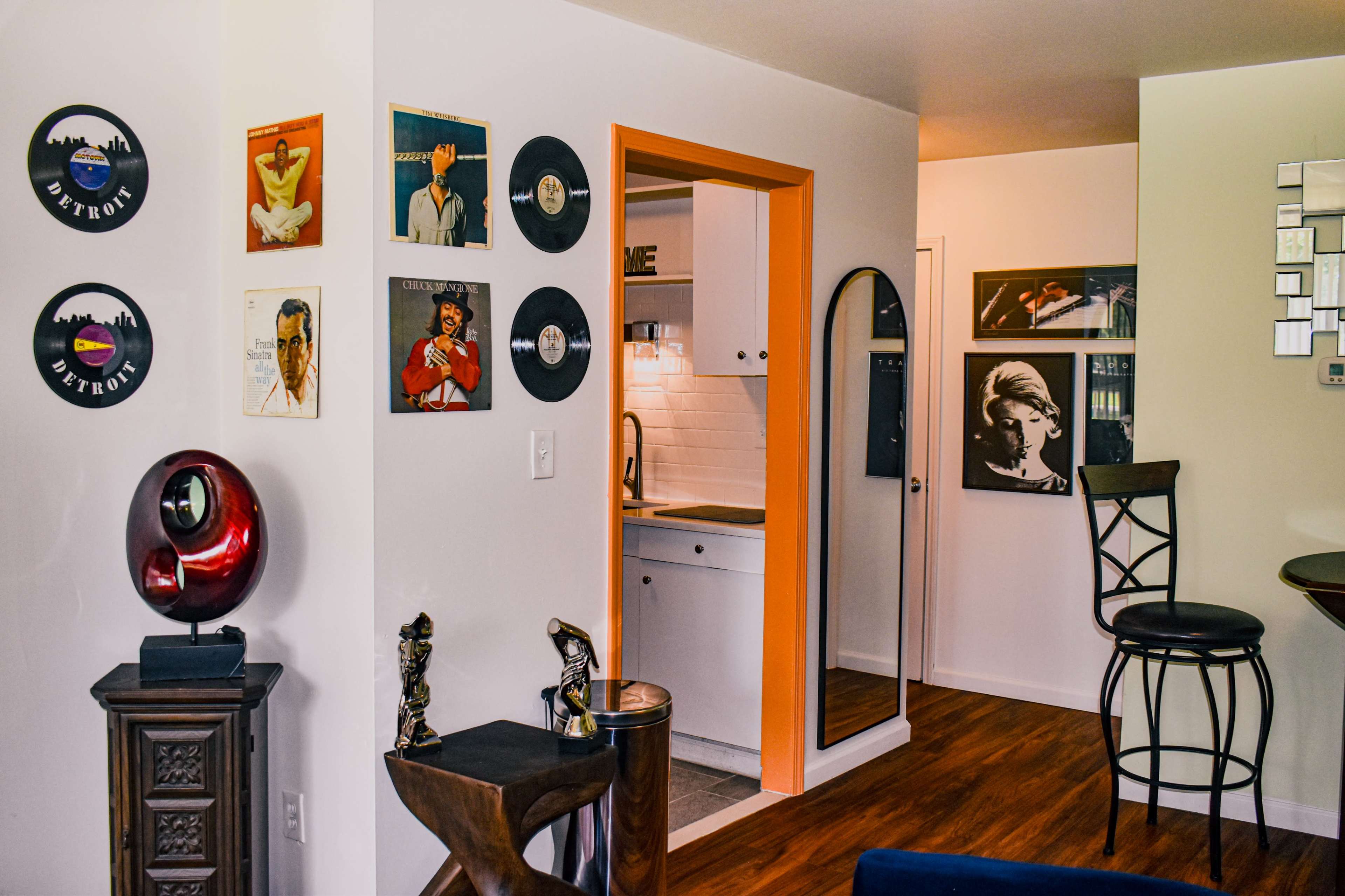The image shows a modern living space featuring framed vinyl records on the wall, a kitchen area in the background, and a bar counter with high stools.