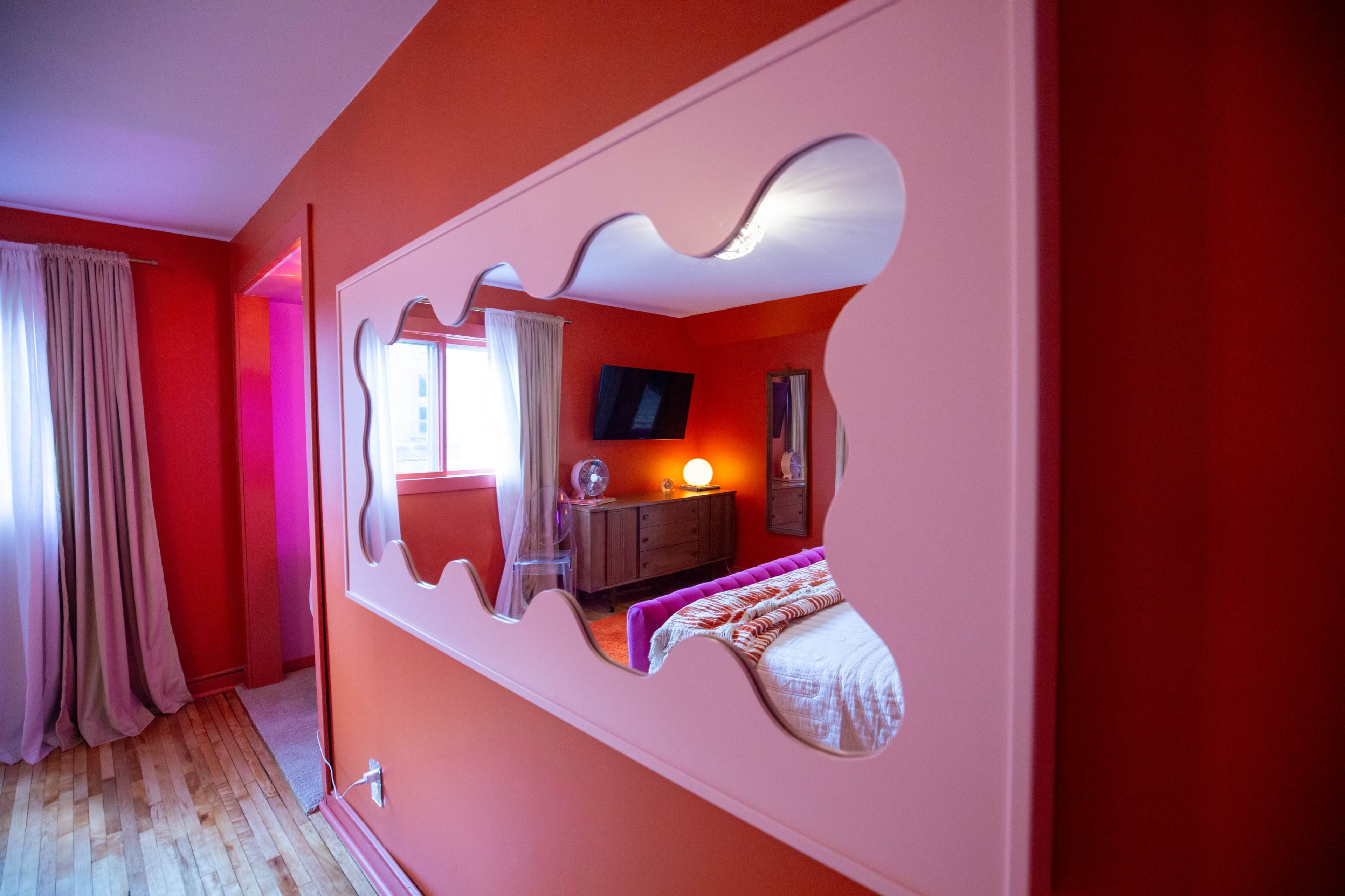 A room featuring a vibrant orange wall and a decorative mirror with a wavy edge reflecting soft curtains and furniture.