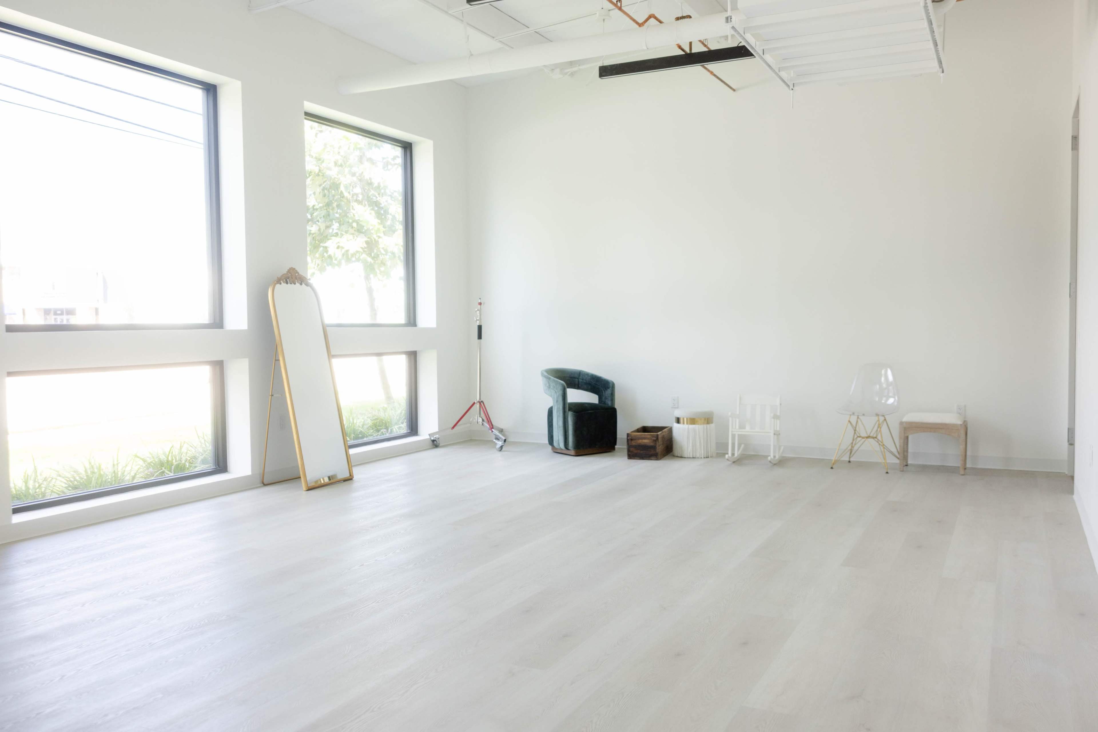 The image shows a bright, minimalist room with two large windows, light wooden flooring, and various pieces of furniture including a mirror and chairs.