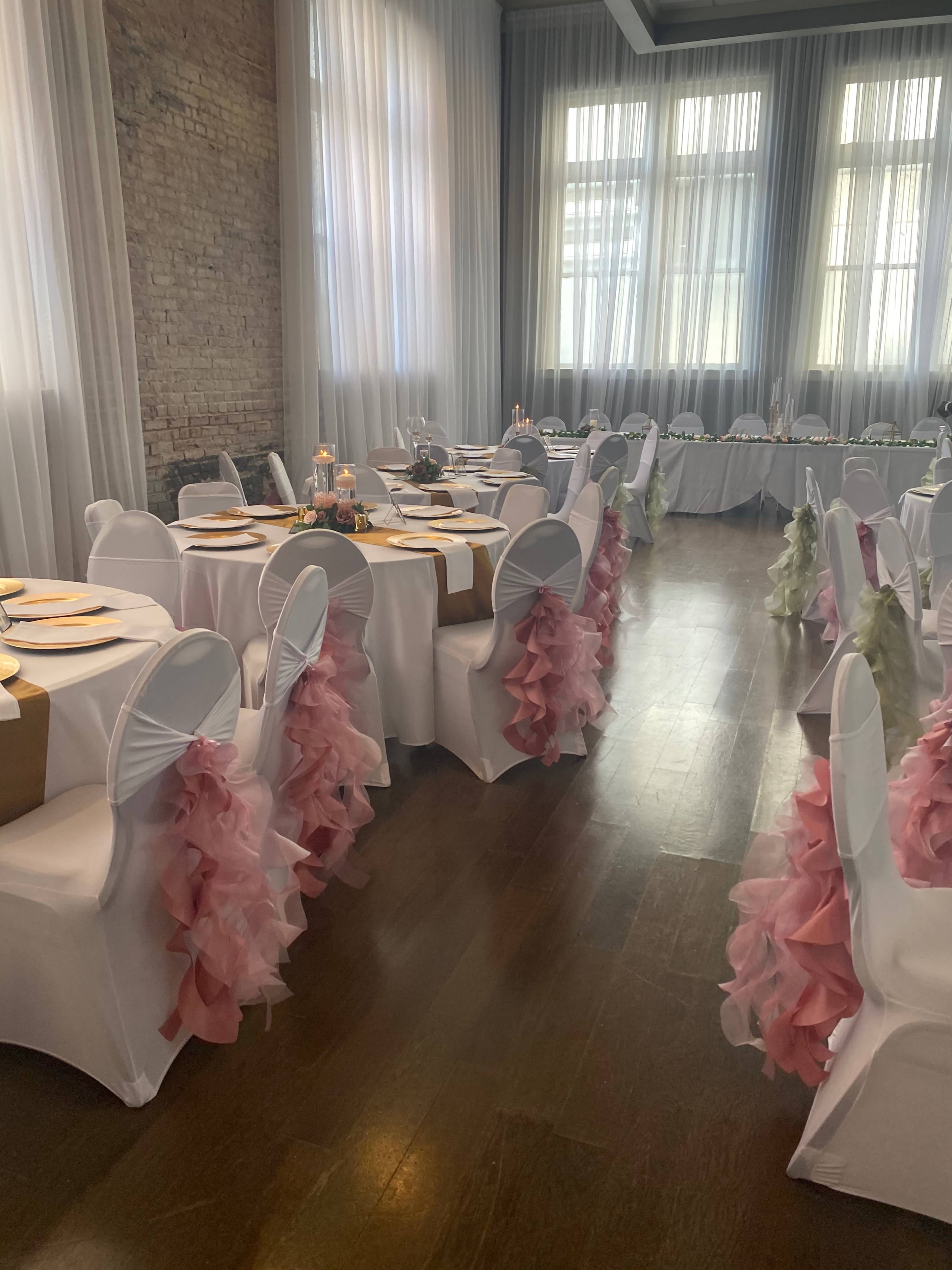 A banquet hall set for an event, featuring tables with white tablecloths and chairs adorned with pink and green ruffled covers, along with elegant centerpieces and candles.