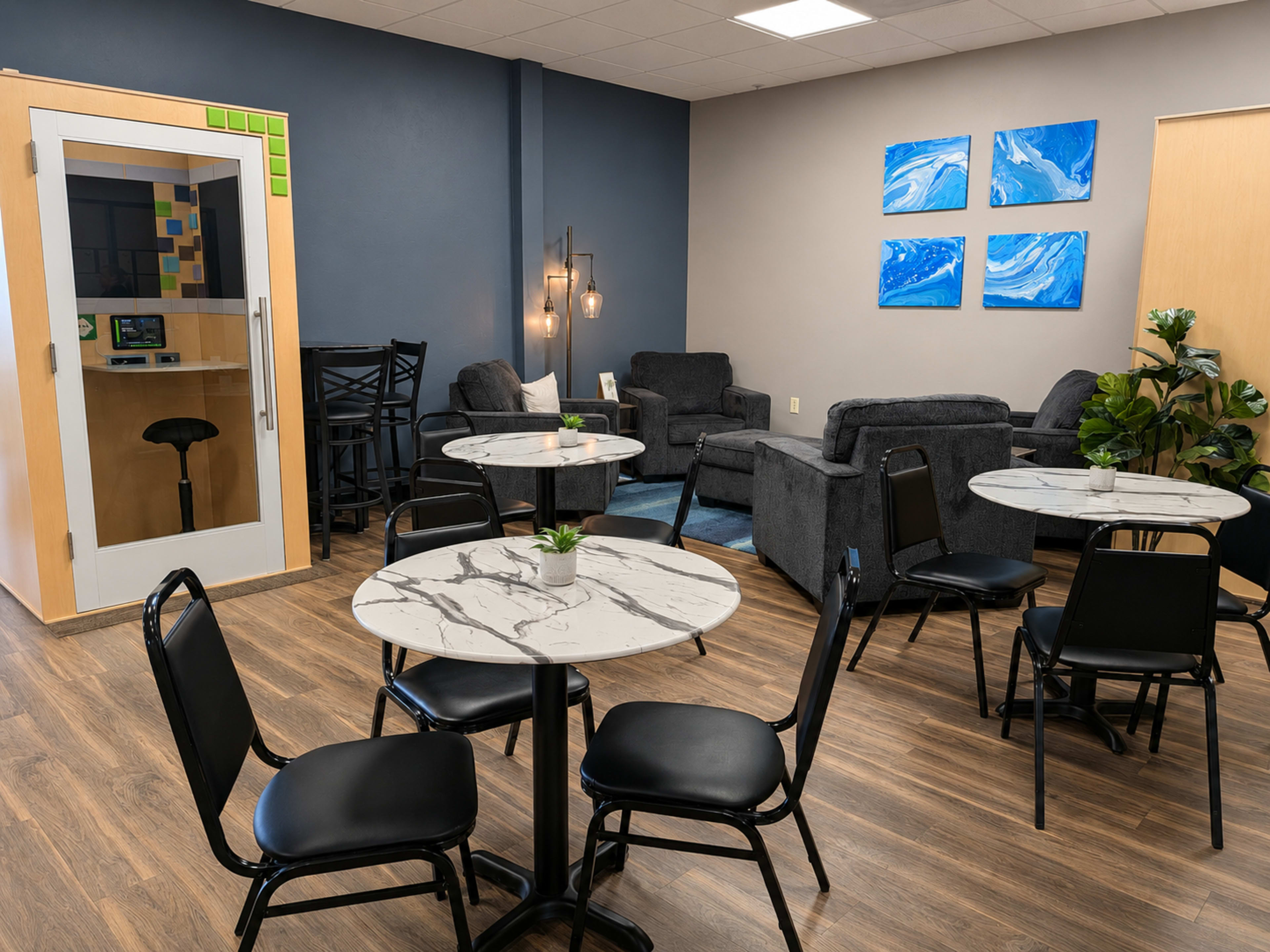 The image shows a modern lounge area with black chairs and marble-top tables, featuring gray sofas, paintings on the walls, and a glass-walled phone booth in one corner.