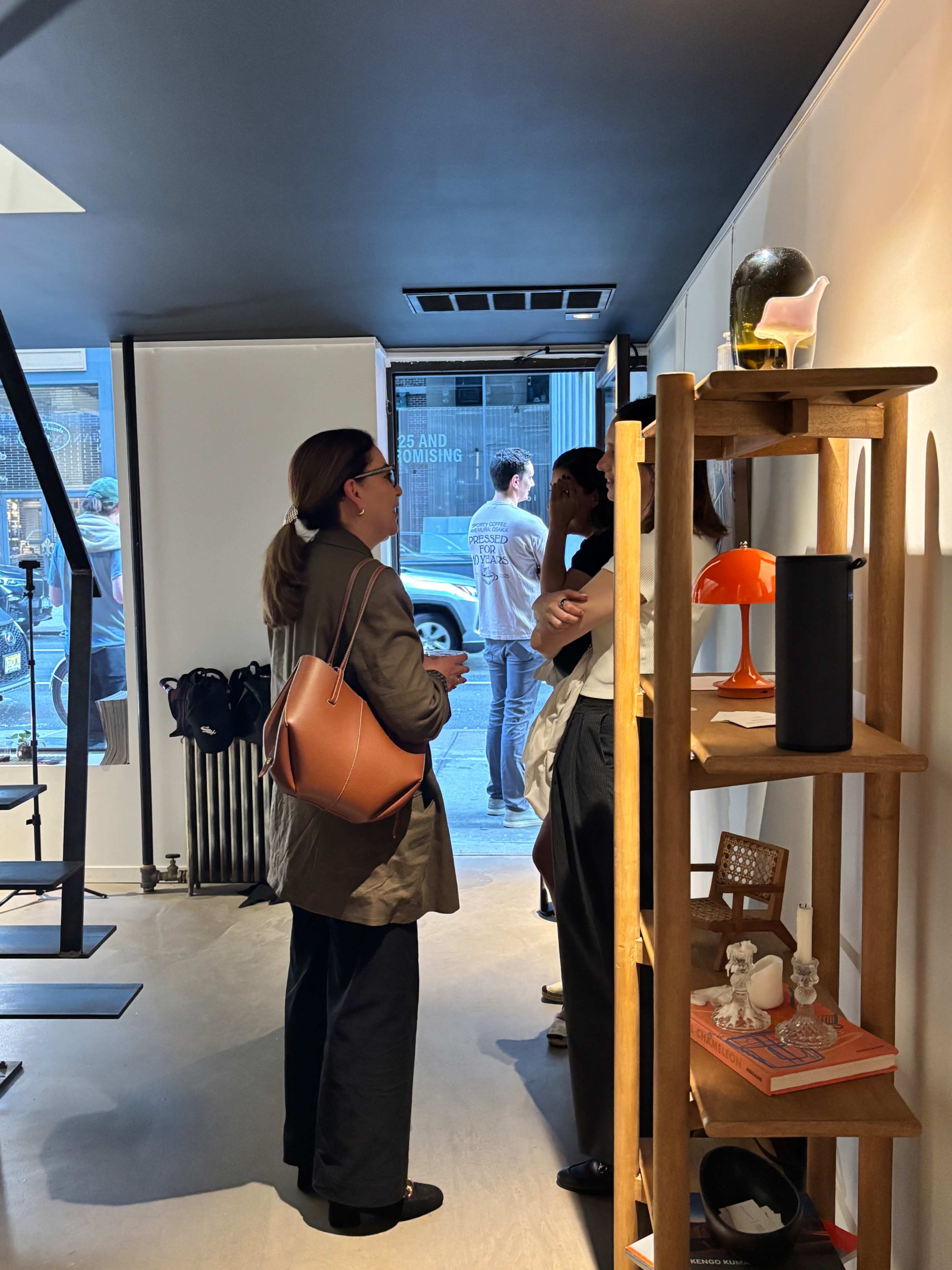 Two women stand conversing inside a shop, while a man is visible outside through the large glass door.