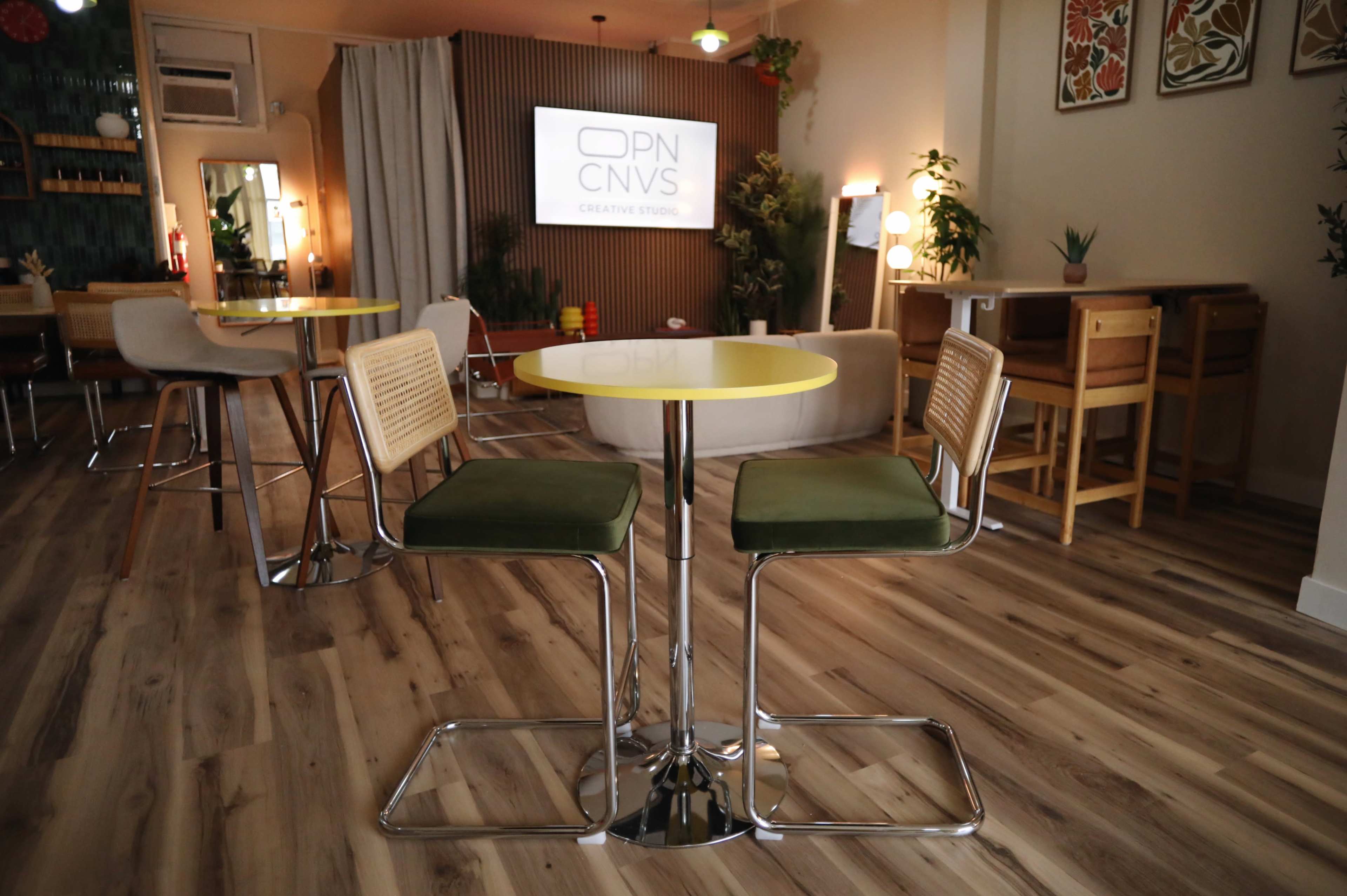 The image shows a modern creative studio space featuring yellow-topped tables with green chairs, wood flooring, and a projection screen displaying "OPN CNVS" on the wall.