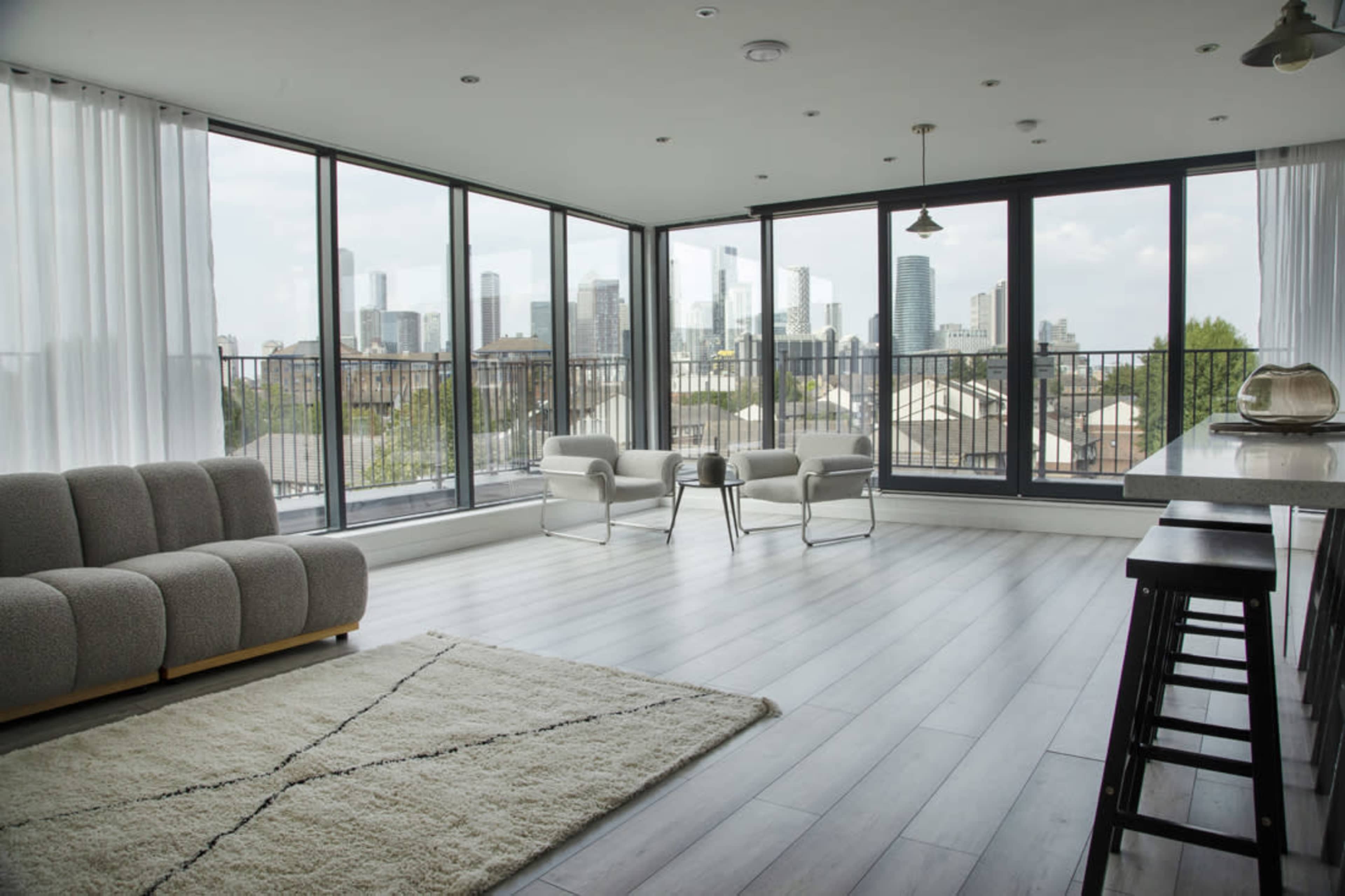 The image shows a modern living space with large windows overlooking a city skyline, featuring a grey sofa, two white chairs, and a light area rug on hardwood flooring.