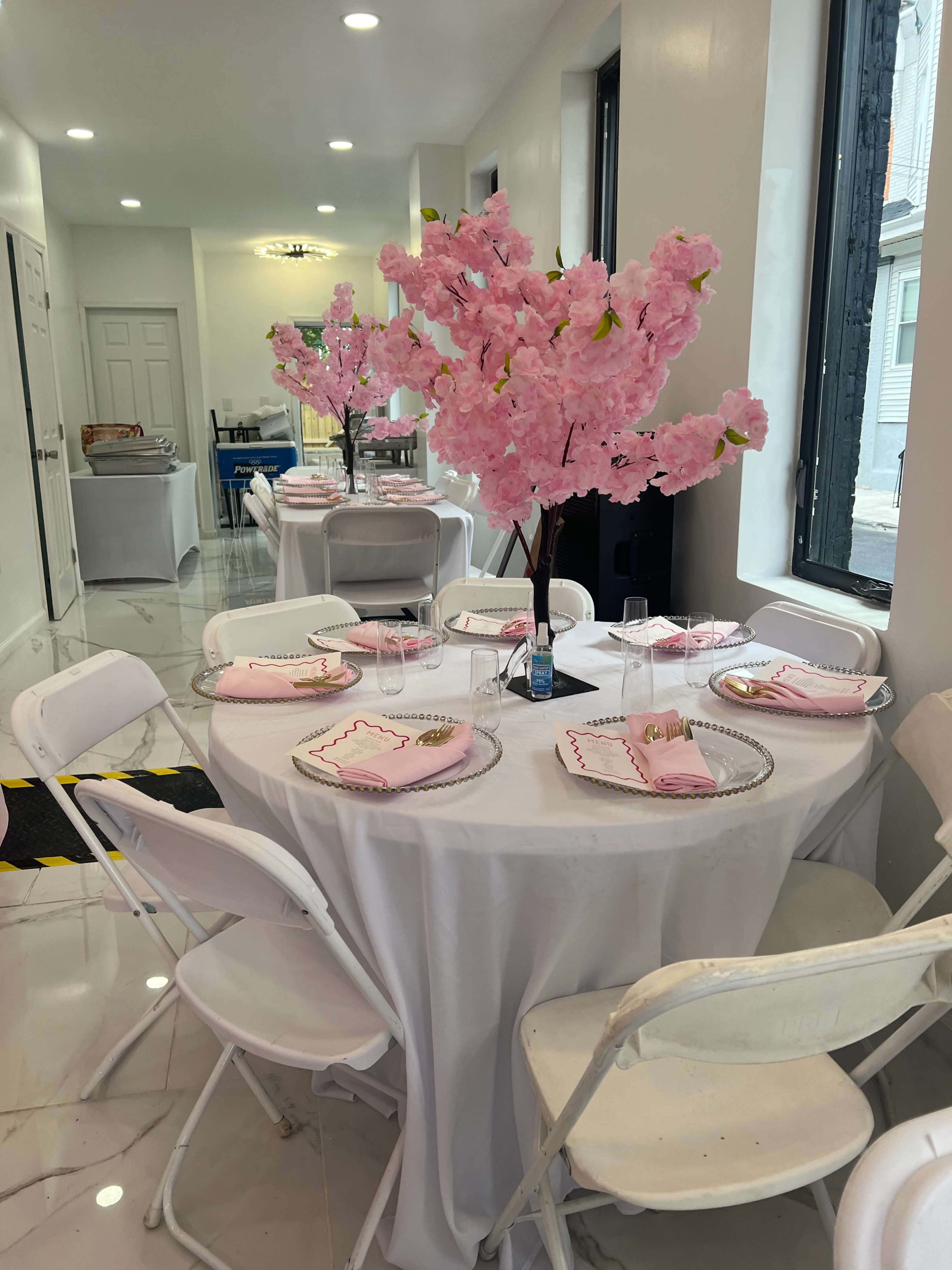 A round table is set for a meal with pink plates and napkins, surrounded by white folding chairs, with a large pink cherry blossom centerpiece.