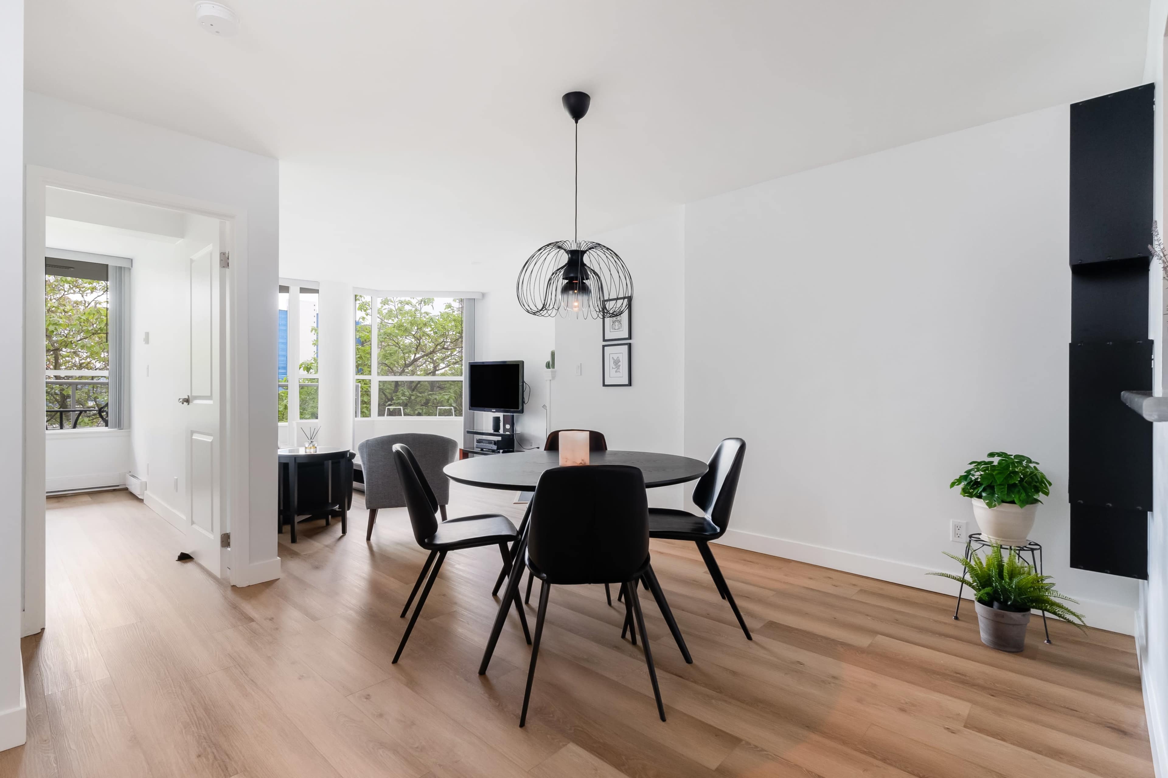 A modern dining area features a round table surrounded by black chairs, with large windows allowing natural light and greenery to view outside.