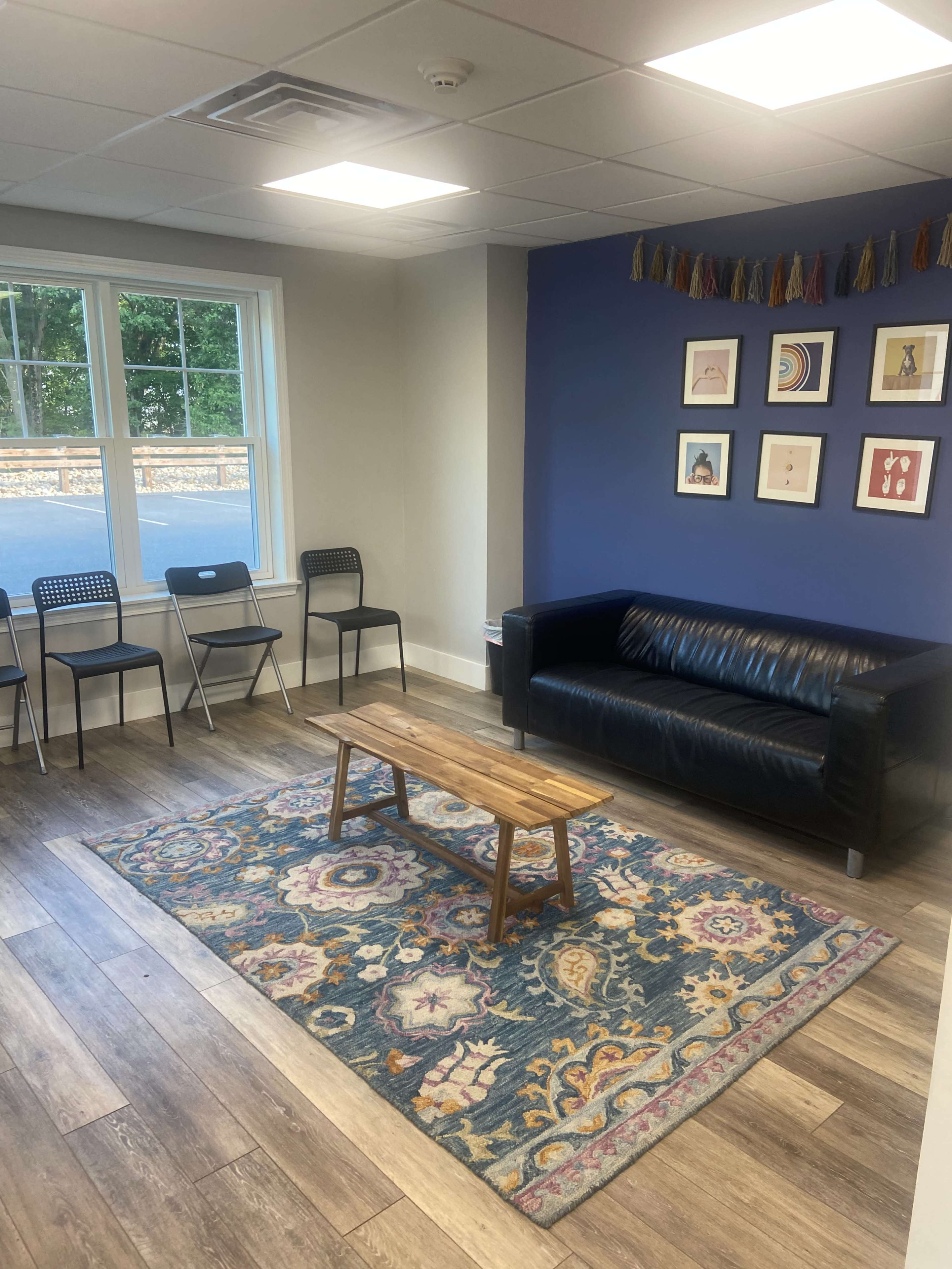 The image shows a modern lounge area with a blue accent wall, a black sofa, a wooden coffee table, patterned rug, and some chairs arranged around the space.