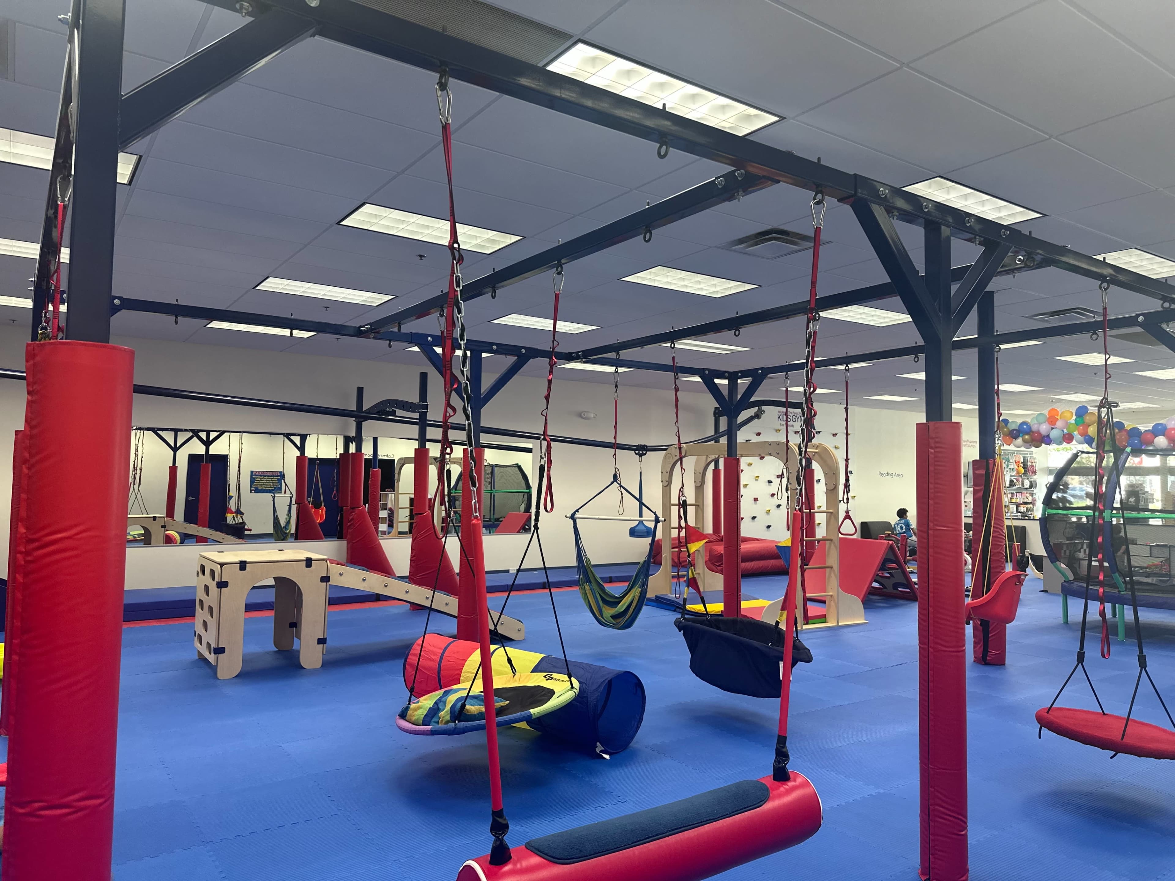 The image shows an indoor play area filled with various colorful equipment, including swings, hammocks, and climbing structures hanging from the ceiling.