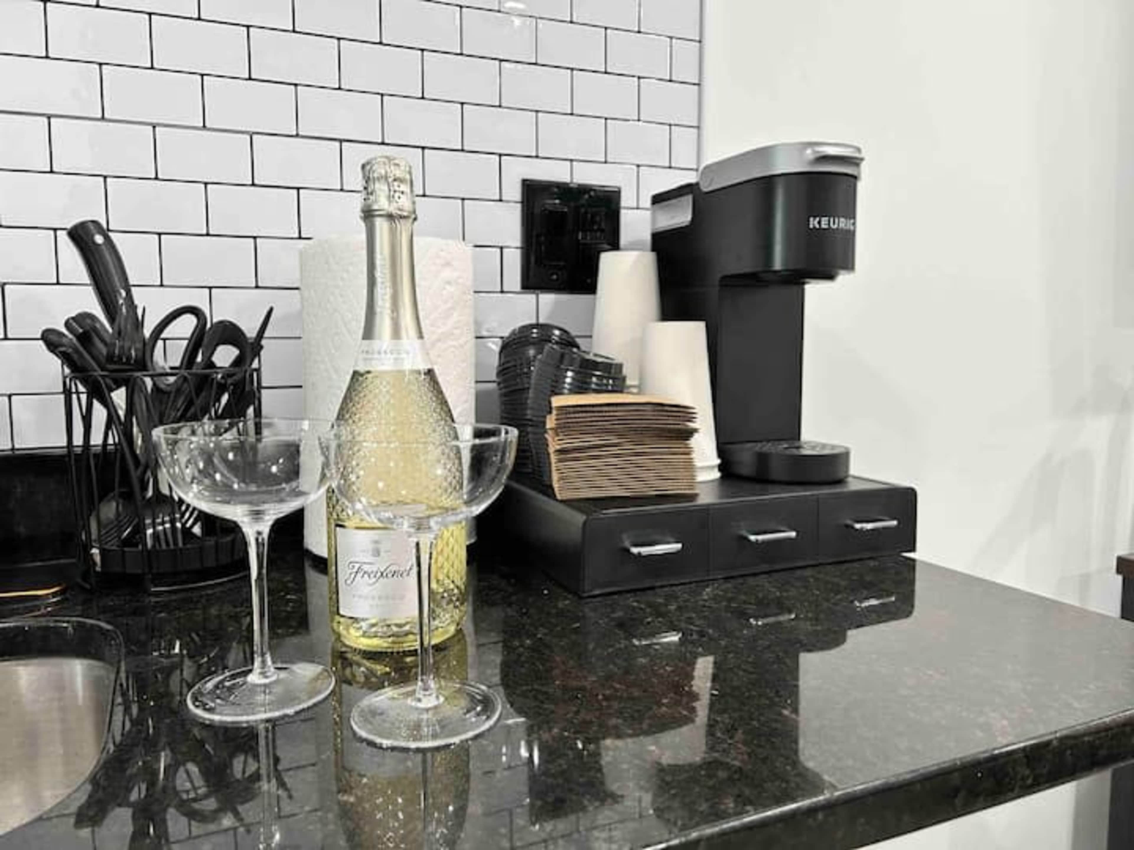 A bottle of sparkling wine stands next to two champagne glasses on a black countertop, with a coffee maker and utensils in the background.