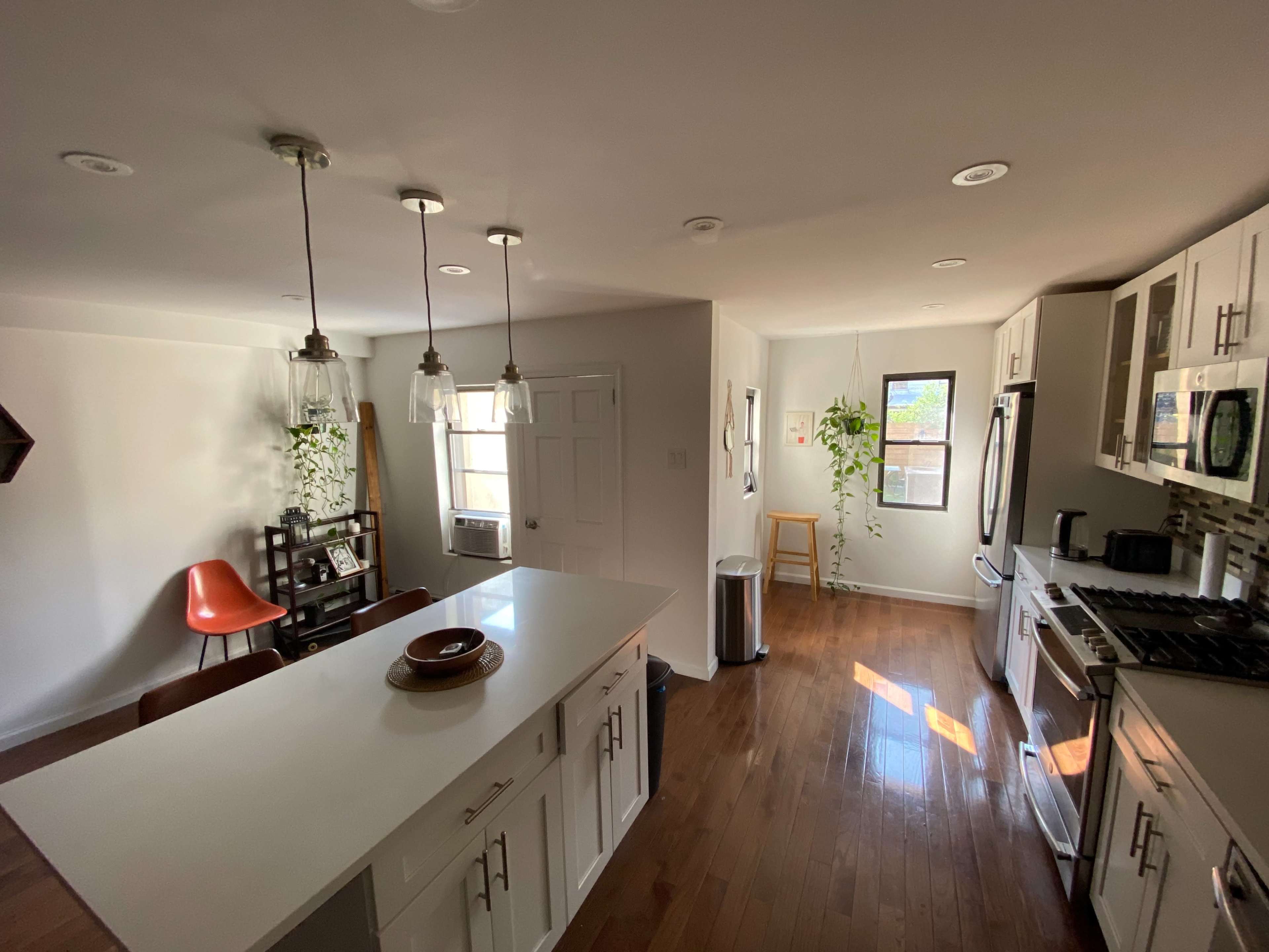 A modern kitchen with a central island, equipped with a stove, refrigerator, and an eating area, alongside an adjacent living space with a chair and a window.