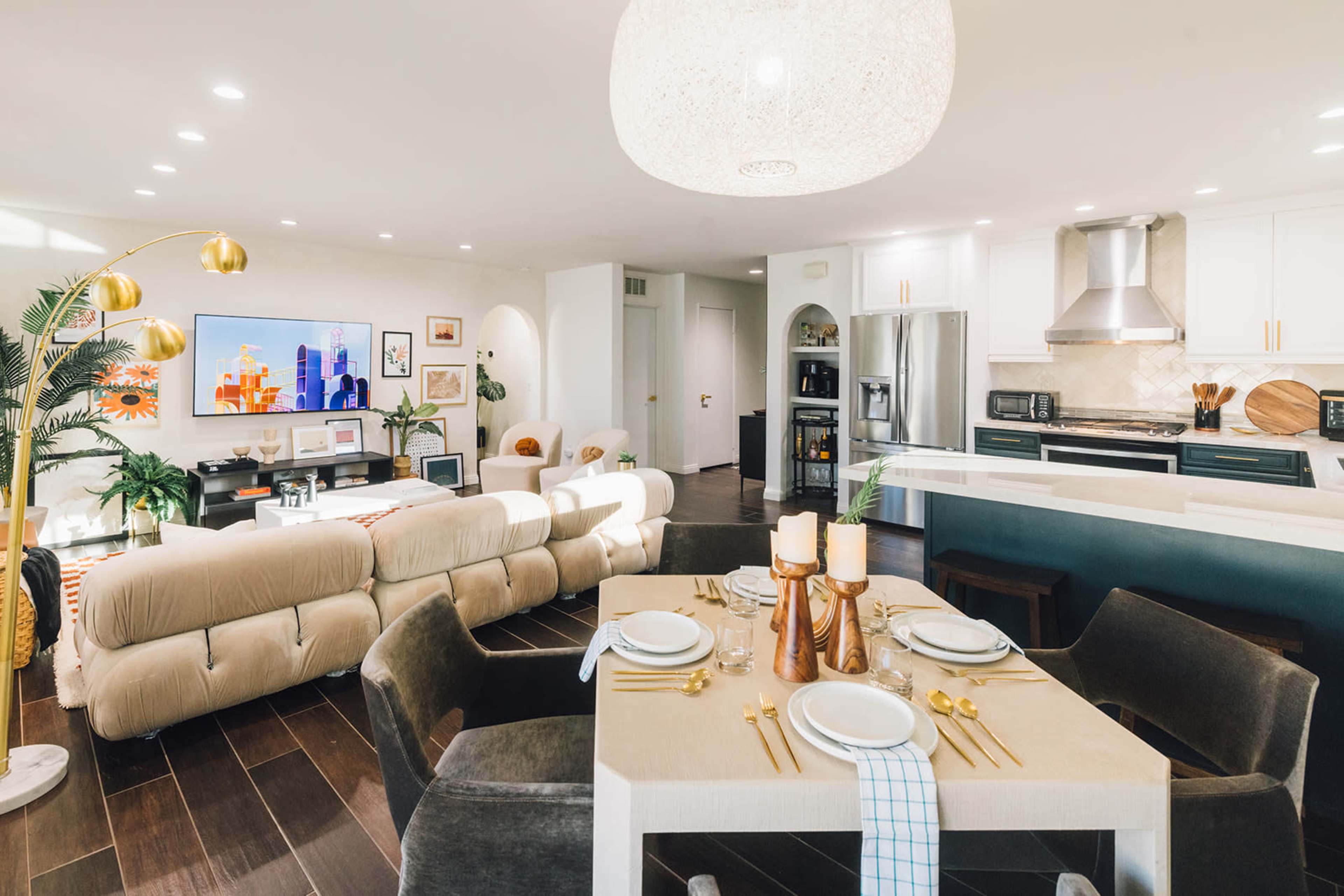 A modern living space features a dining table set for four, a sectional sofa, a large TV, and an open kitchen area with white cabinetry.