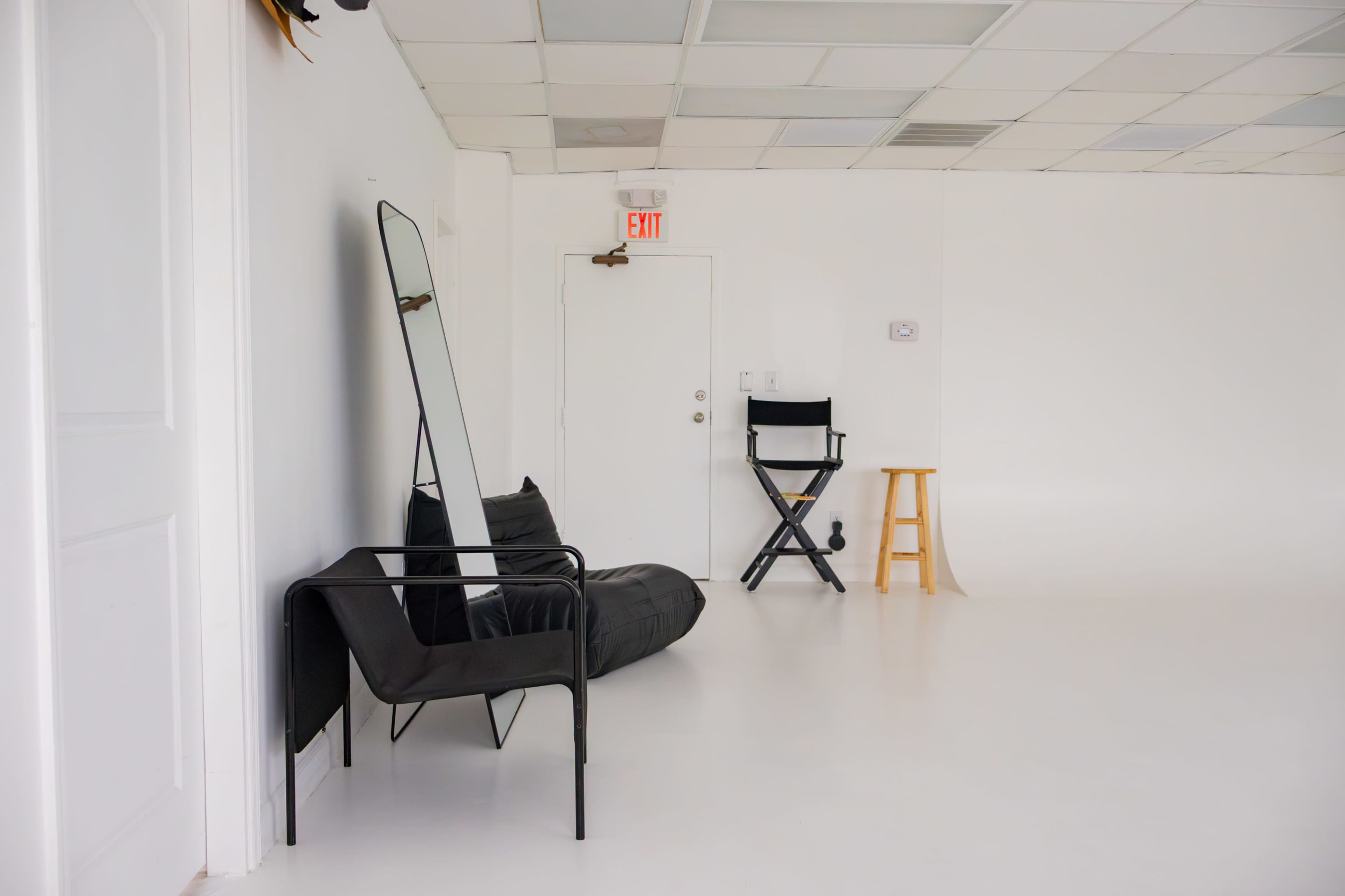 The image shows a minimalist, white interior space featuring a black chair and beanbag, a tall mirror, a director's chair, and a wooden stool.