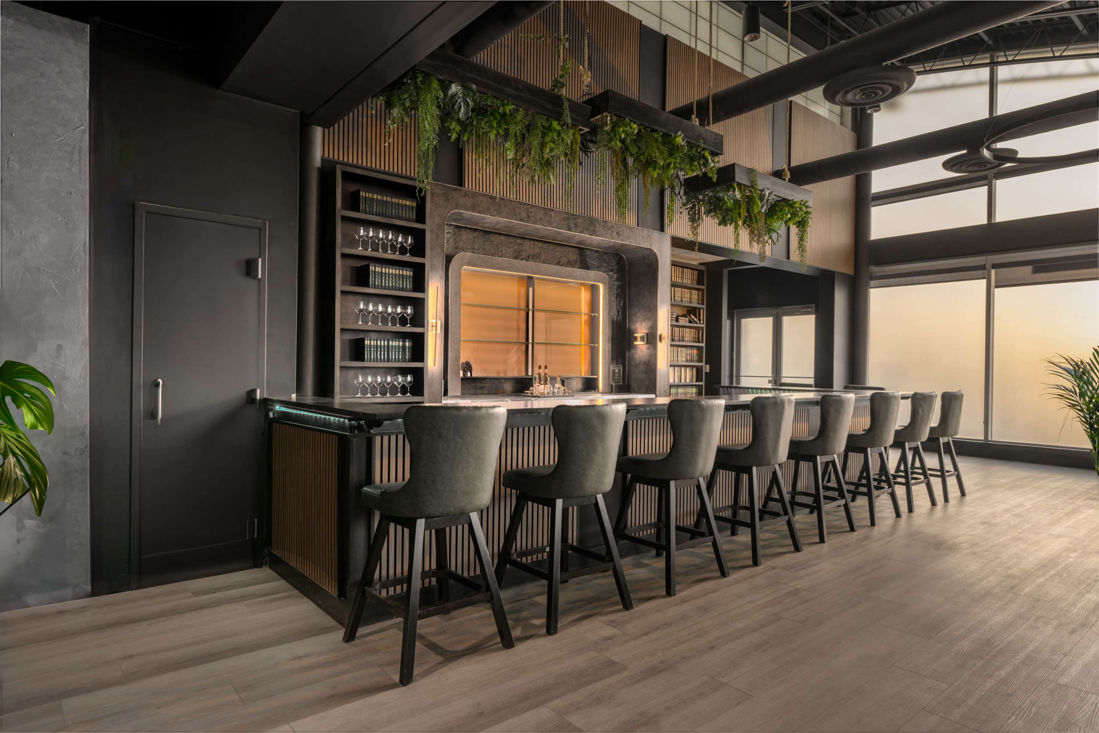 A modern bar with a sleek design, featuring a long counter, high stools, and greenery hanging from the ceiling.