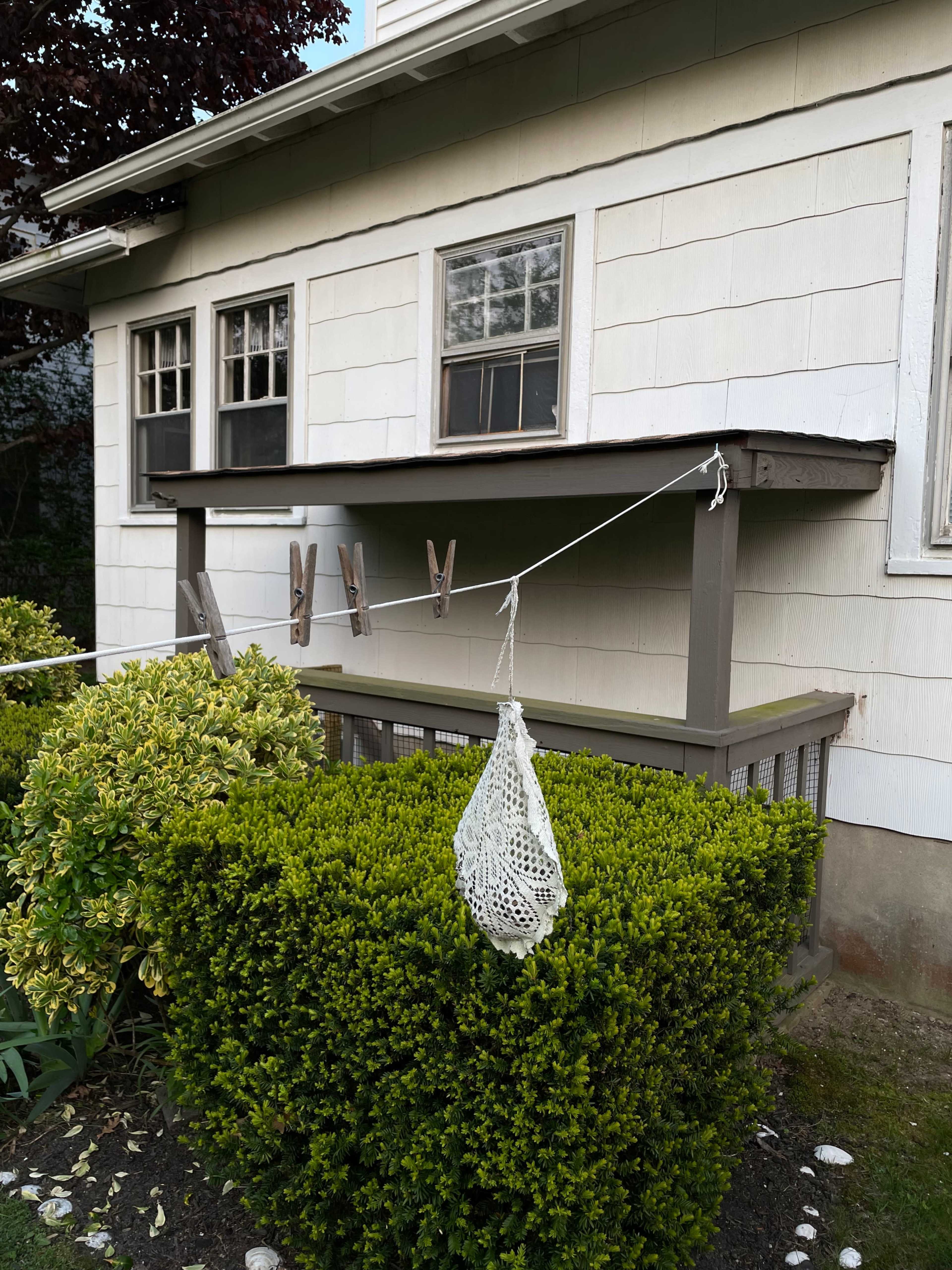 A clothesline with wooden clothespins and a hanging crochet bag is attached to a porch beside a neatly trimmed hedge in front of a house.