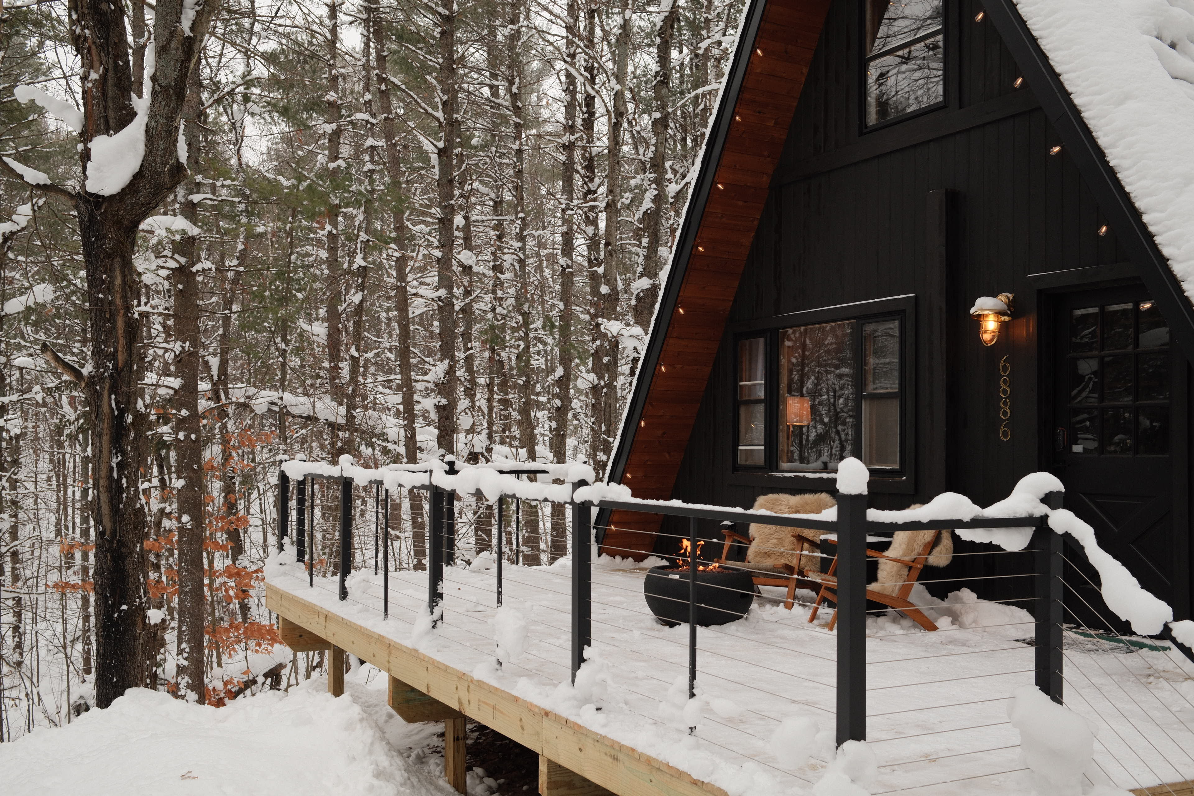 A black A-frame cabin with a snow-covered deck is surrounded by tall trees in a winter landscape.