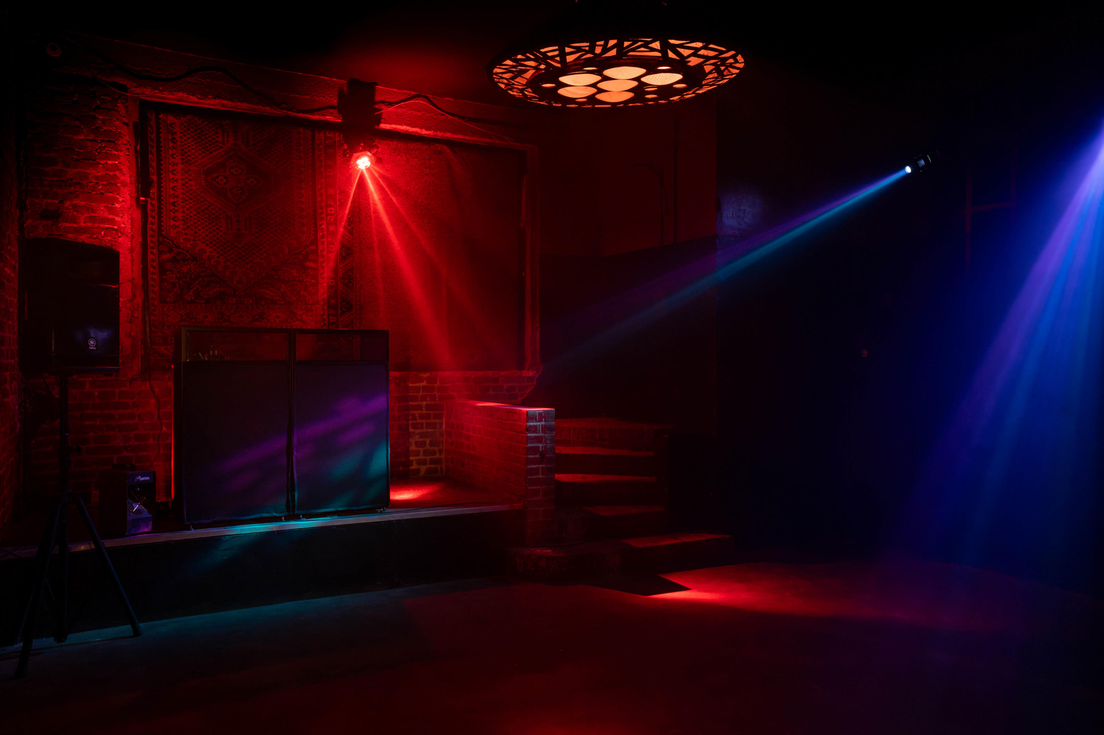The scene features a dimly lit room with vibrant red and blue lighting, a raised platform with speakers, and a decorative wall hanging.