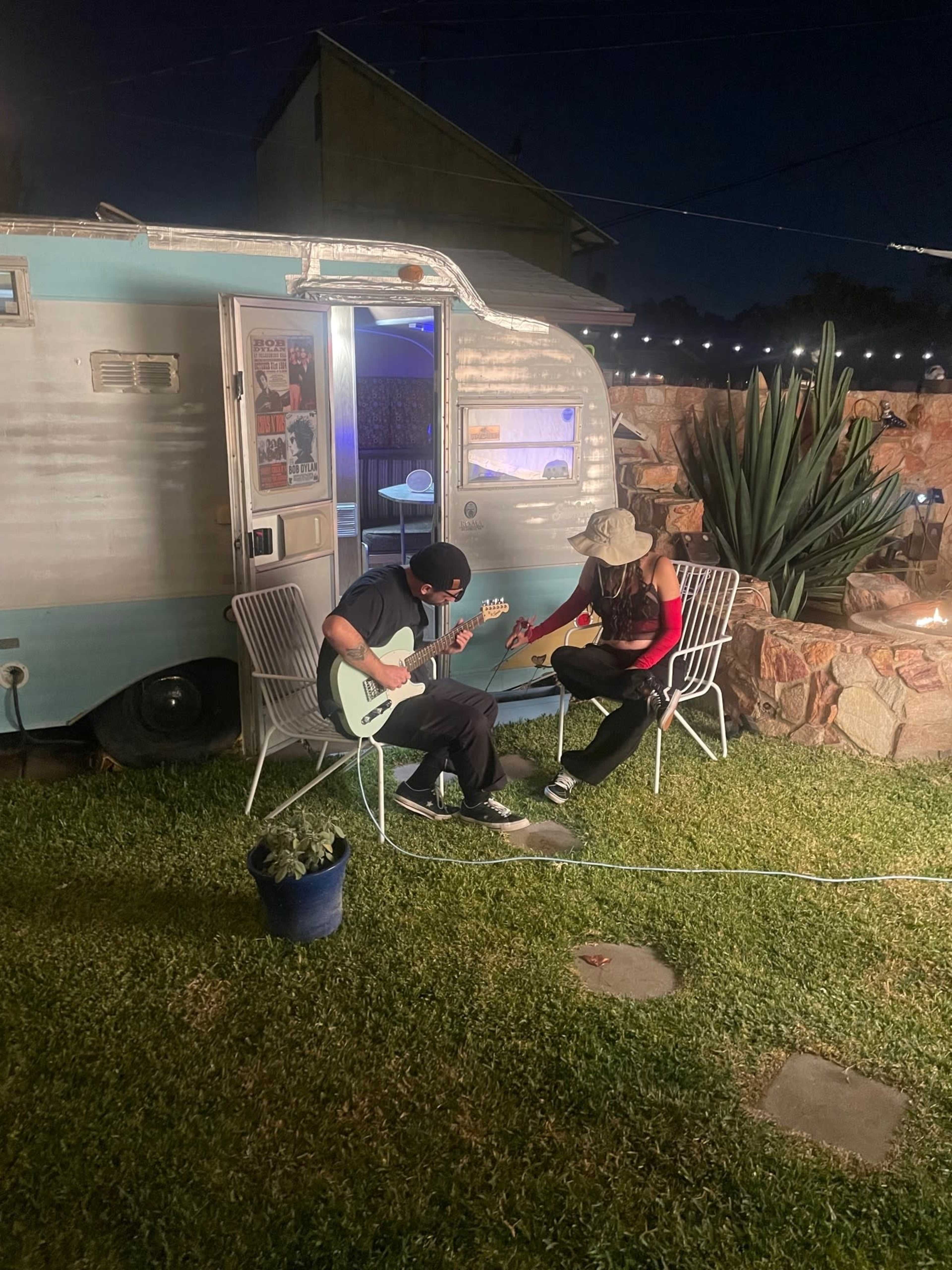 A person plays an electric guitar while another person sits nearby, both enjoying the evening in front of a vintage camper.