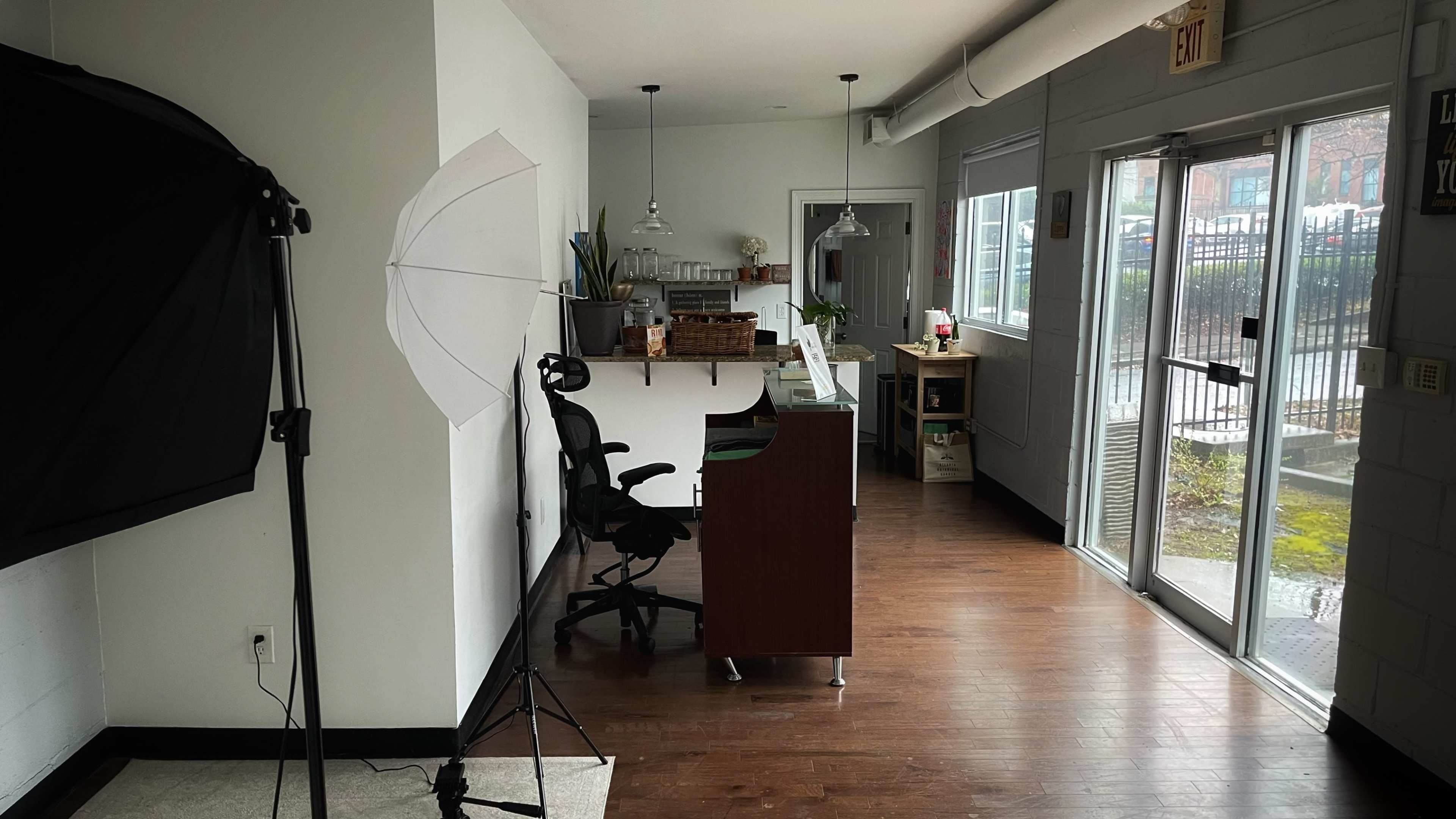 A modern office space with a desk and chair, lighting equipment set up in one corner, and large windows allowing natural light to enter.